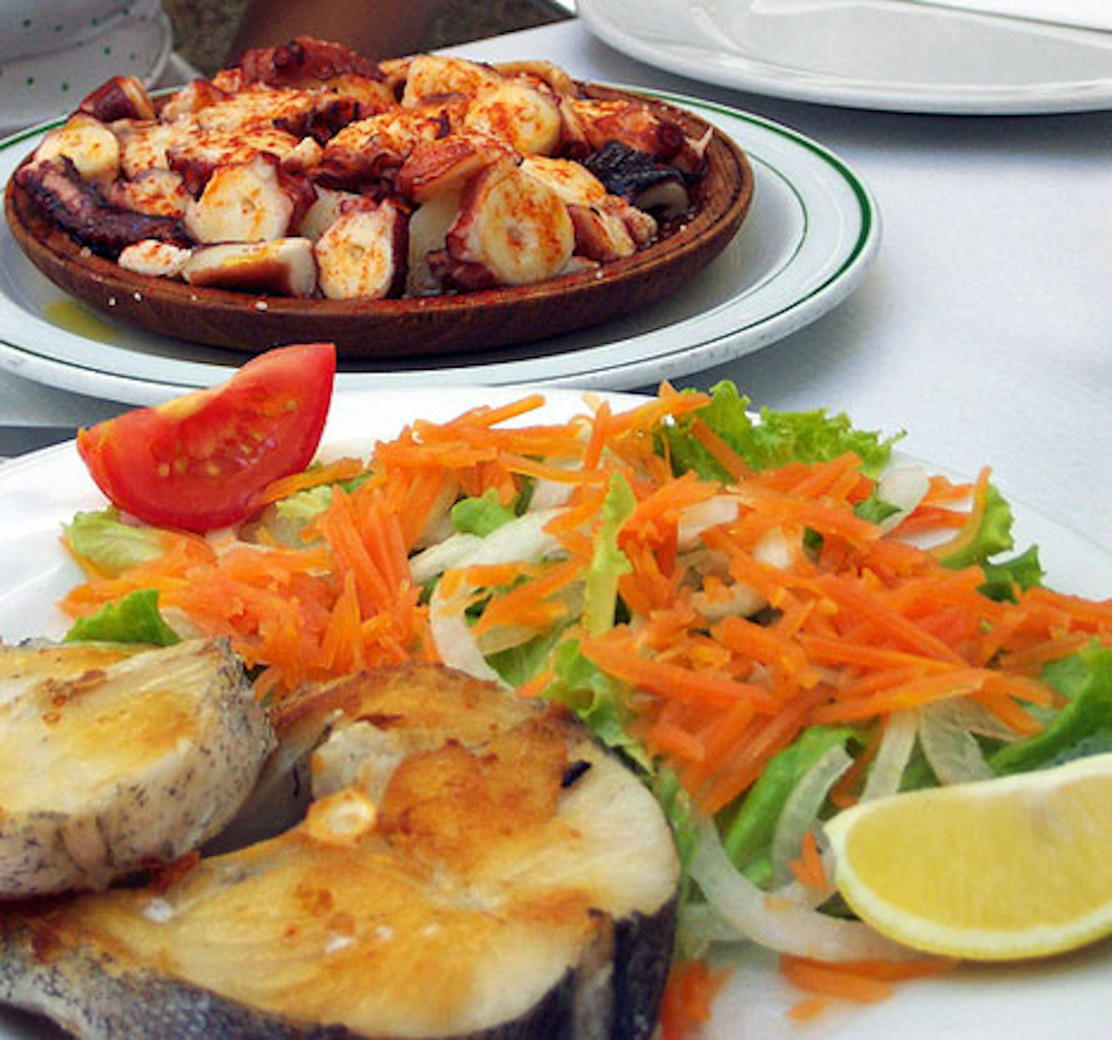 A plate of fried fish, salad and lemon slices in the foreground, and a dark brown platter of paprika-sprinkled boiled octopus in the background