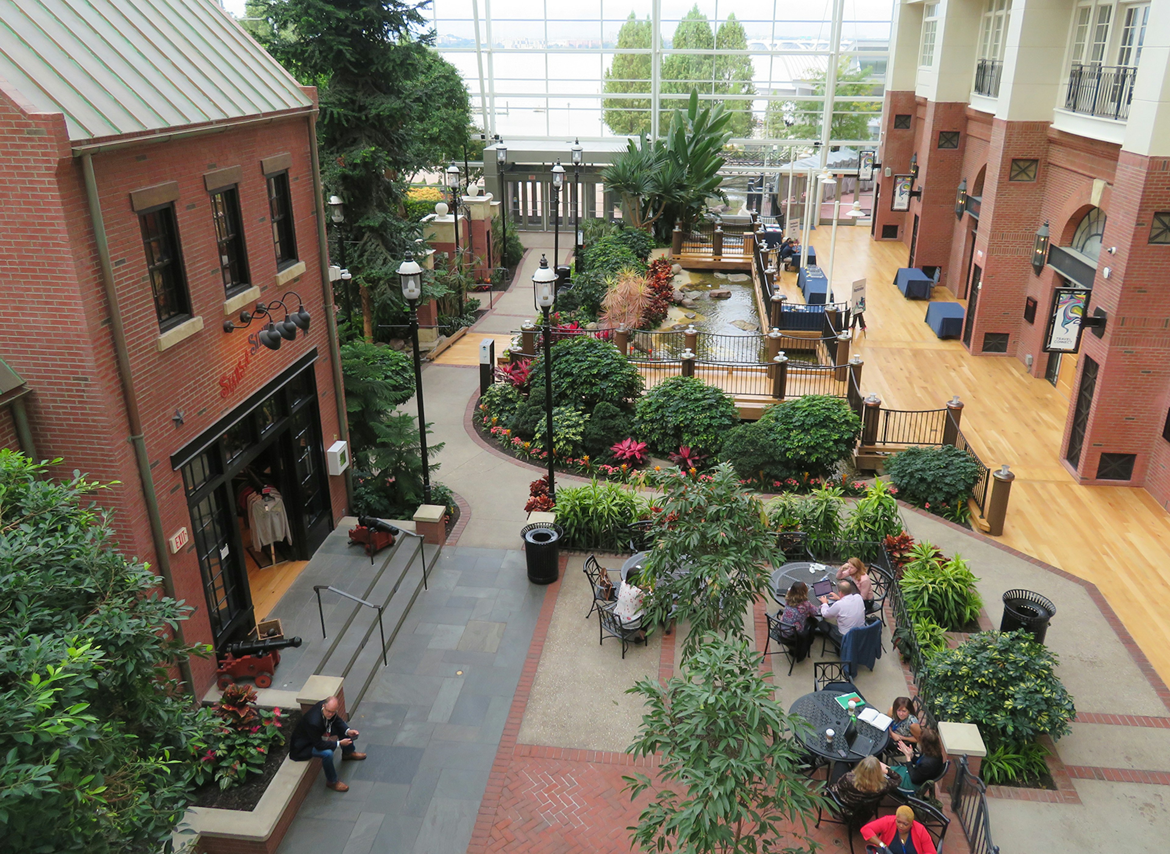 View of the interior of Gaylord National Resort, with brick buildings and tree- and bush-lined pathways under a glass atrium and a few people sitting at wrought-iron tables