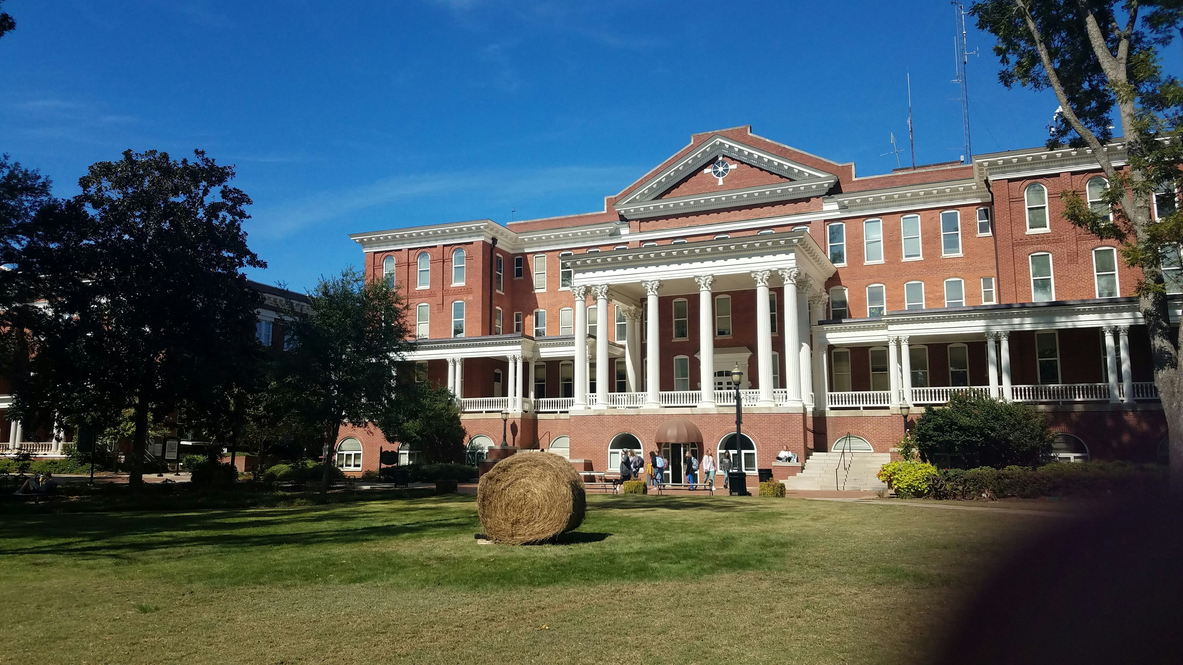 The red brick exterior of a building at the Georgia College and State University, with six white corinthian columns forming a central two-story porch flanked by a lower one-story porch on either side supported by three sets of two columns. A large round hay bale sits on the lawn in the foreground