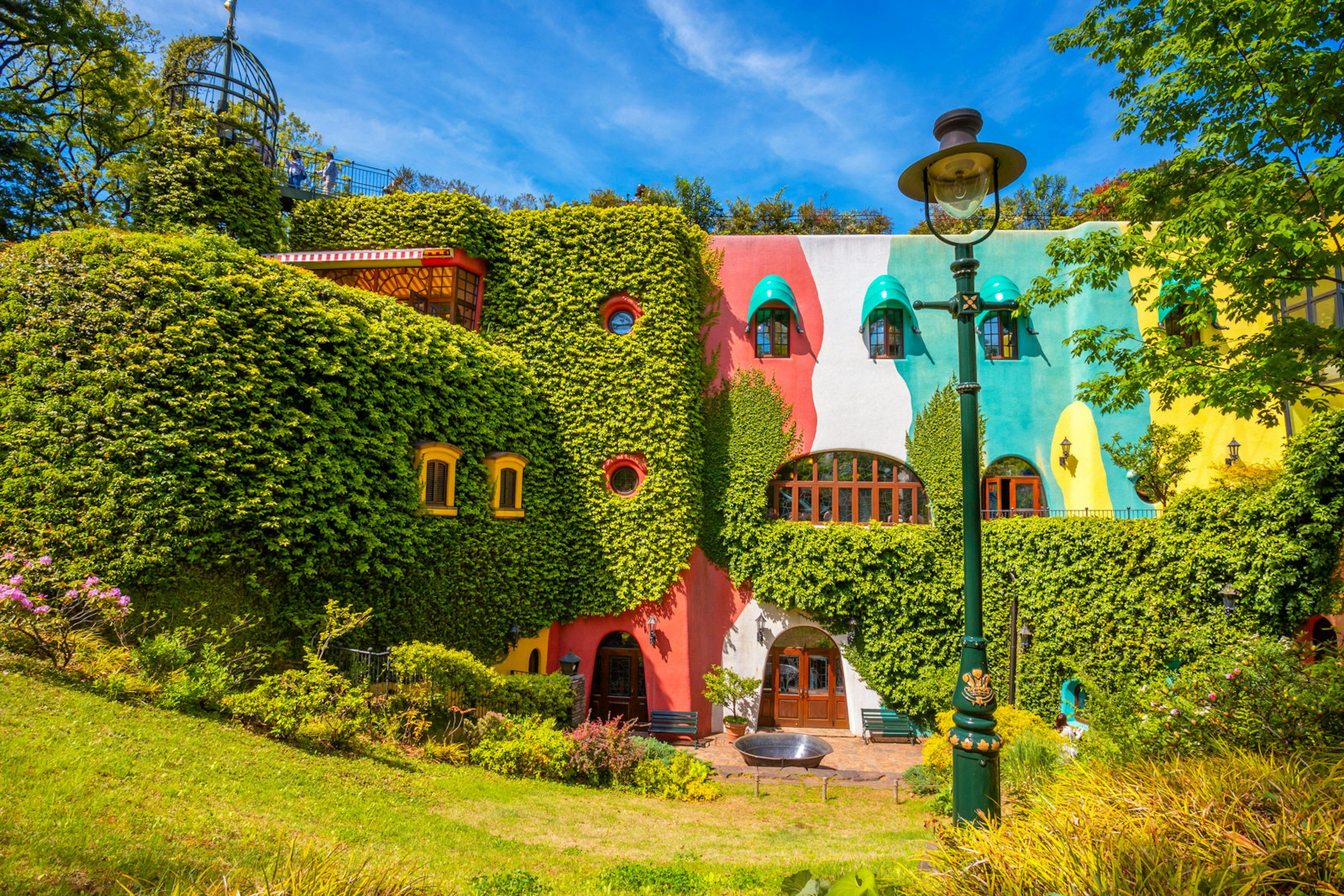 The foliage-clad, brightly coloured exterior of the Ghibli Museum in Tokyo, Japan. The museum is almost completely covered in ivy, the walls that are visible are painted red, white, green and yellow. The windows also have green shades over them and there is a green lamp post in the foreground.