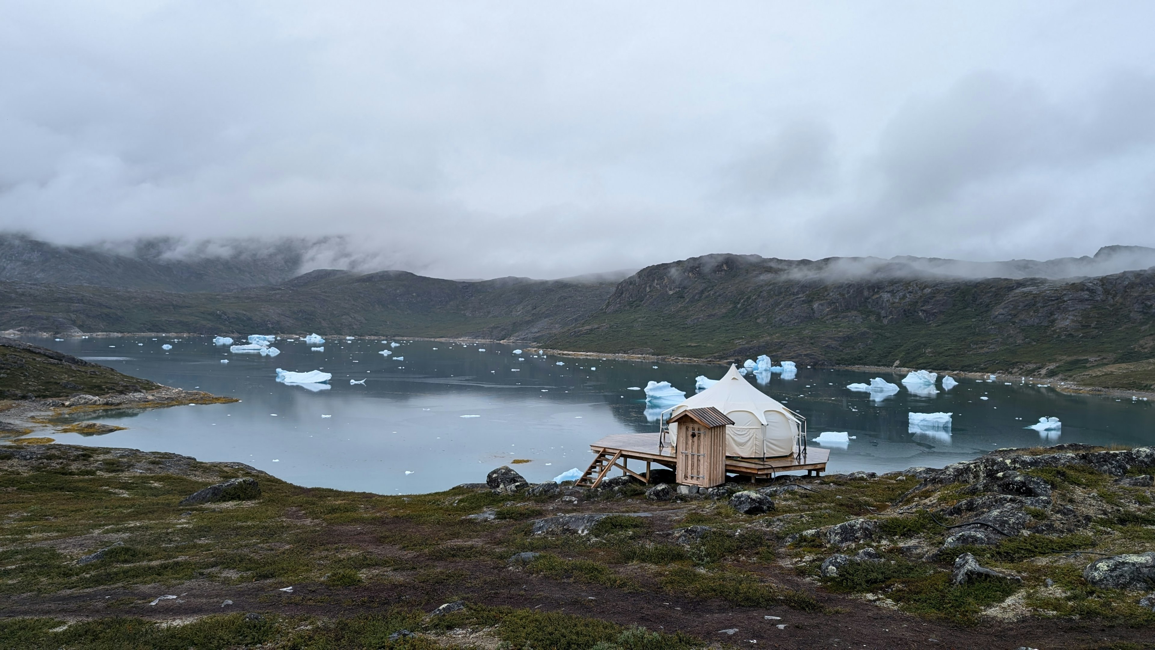 A bell tent and small wooden toilet hut at the edge of a fjord