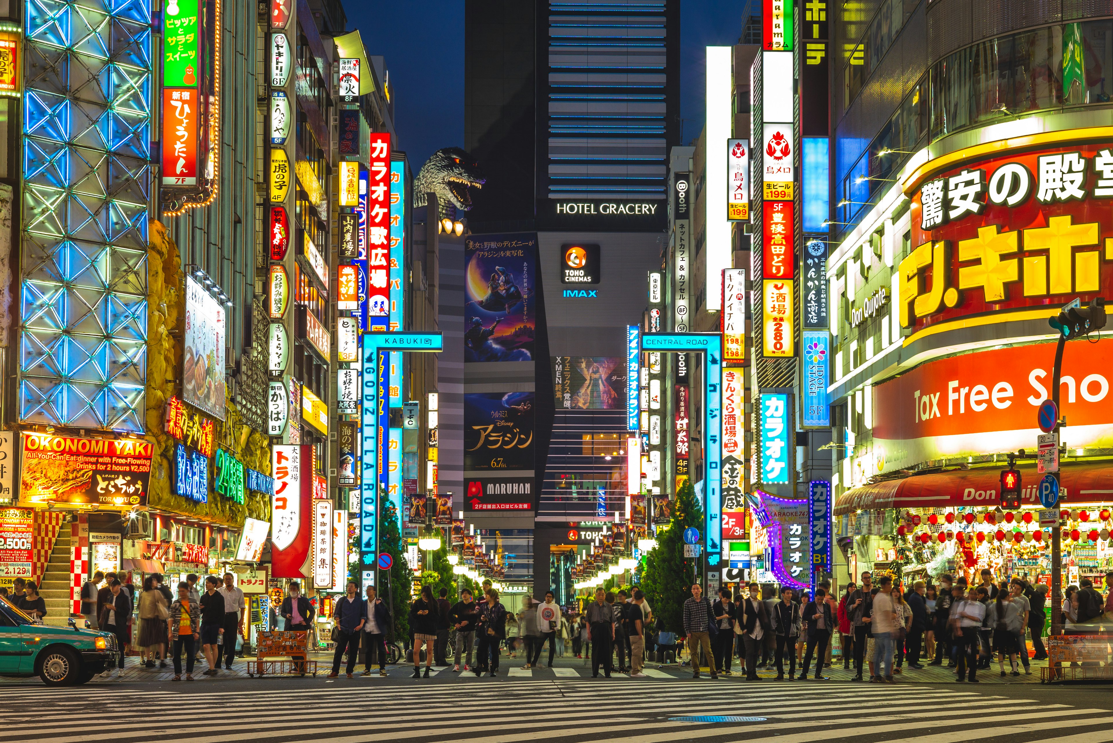 A shot looking down Godzilla Road in Shinjuku, Tokyo; the Hotel Gracery stands at the end of the neon-lit road, and a huge model of Godzilla is looking over a tall building towards it.