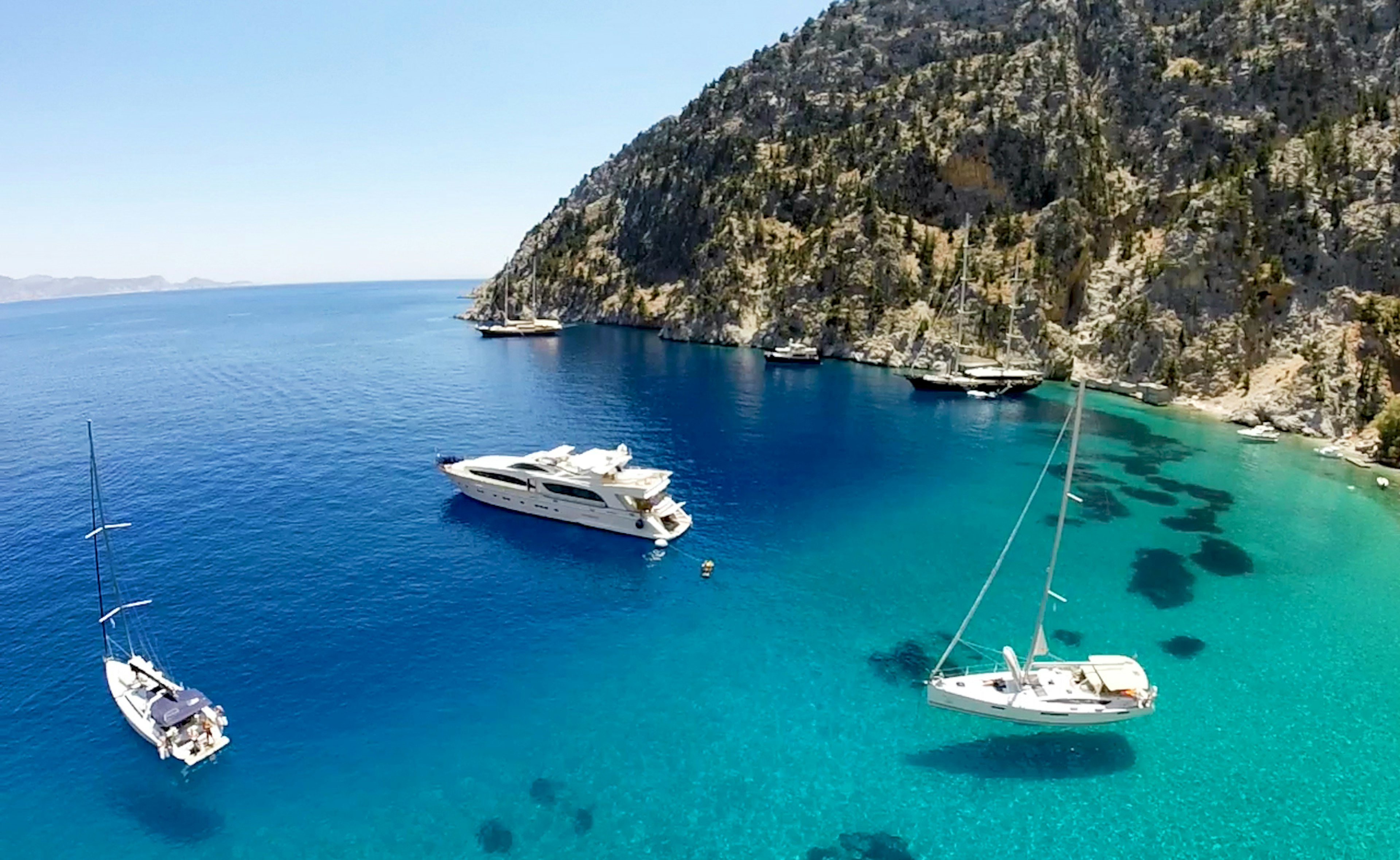 Several white, wedge-shaped boats, one a yacht, one a sail boat, and several too far in the distance to quite make out, sit on deep blue and turquoise waters off the rocky, hilly shores of Greece, which are light brown and dotted with scraggly scrub trees.