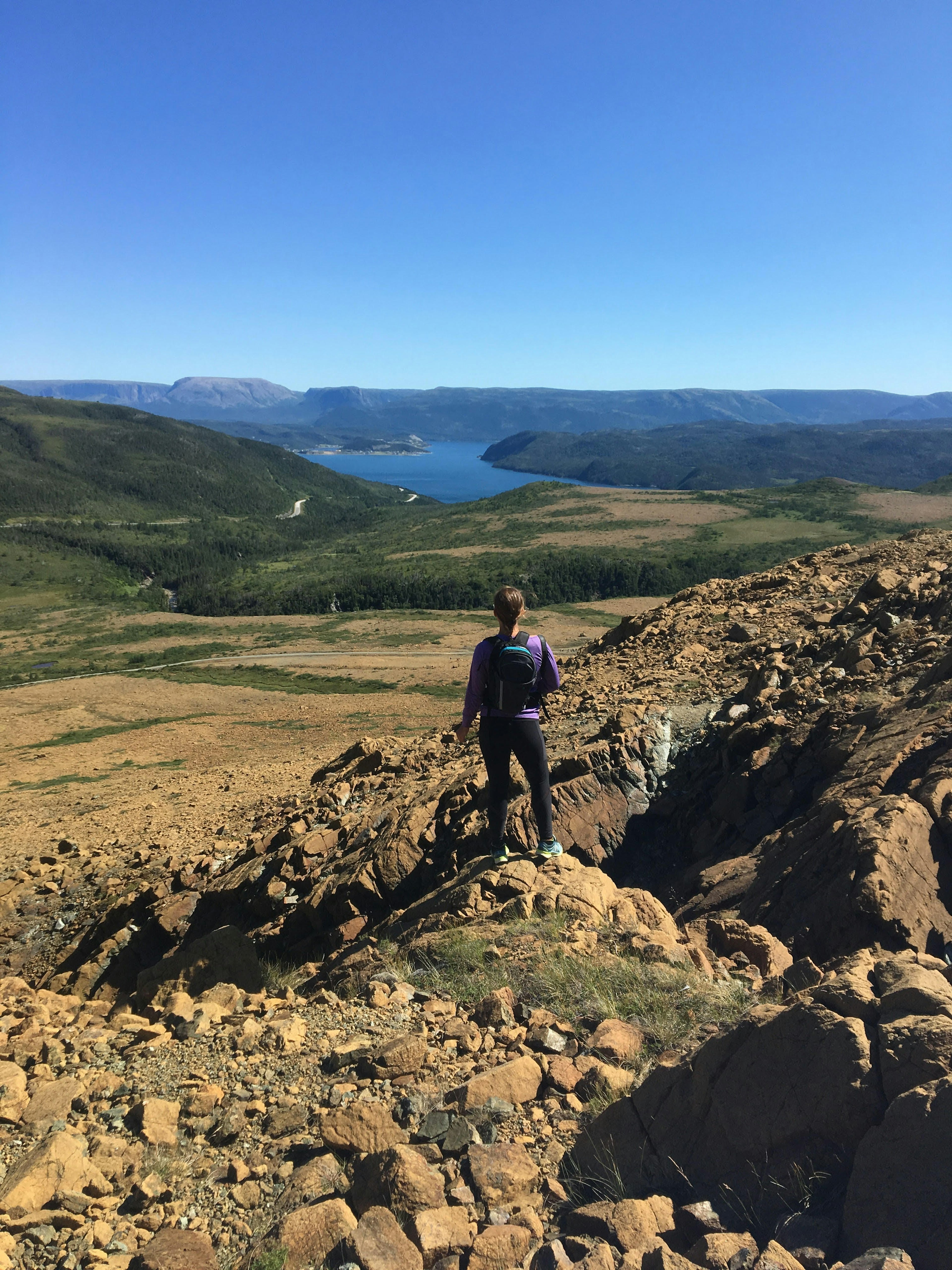 A woman stands on a rocky outcrop with her back to the camera; she is looking down towards a lake in a valley.