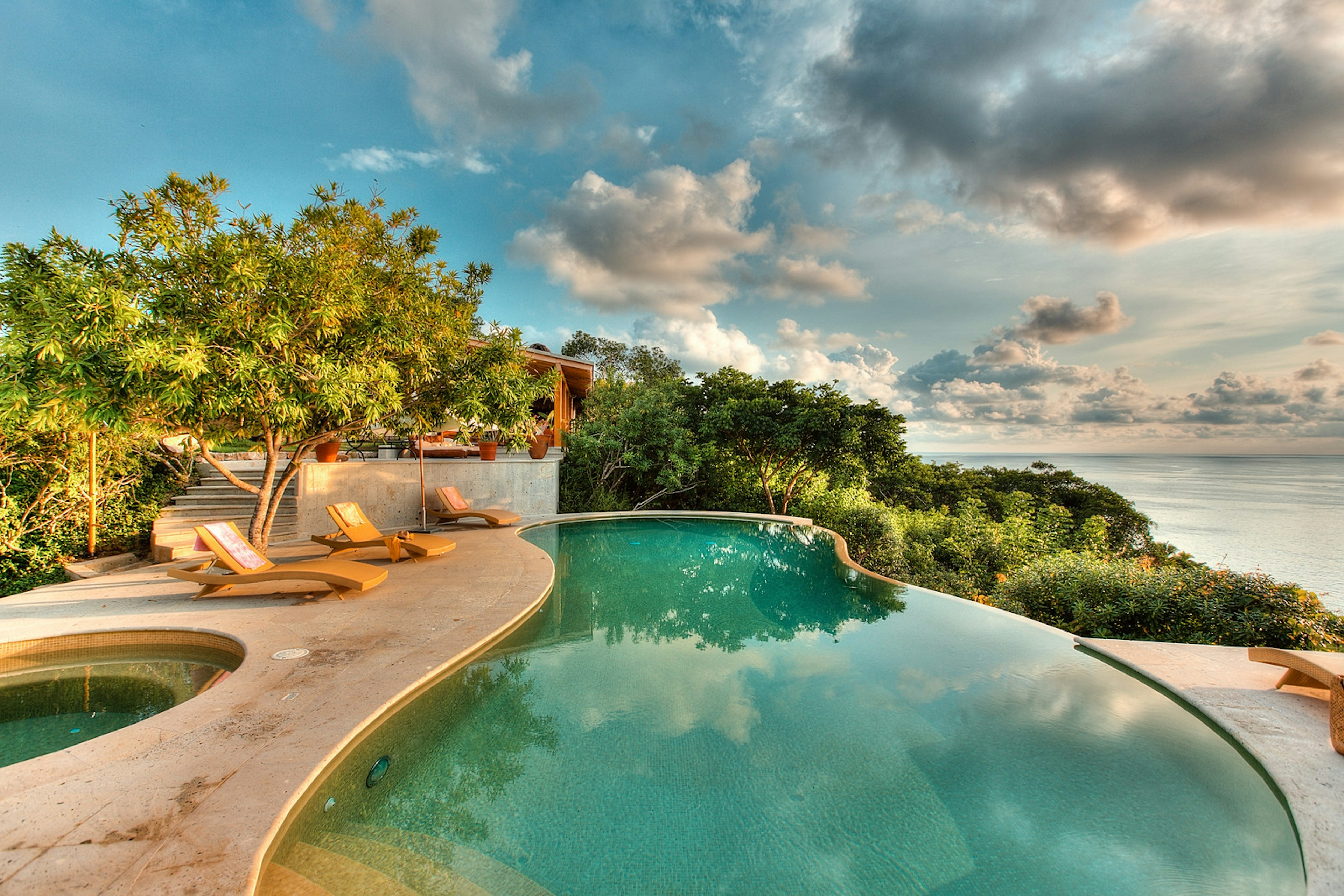 A luxurious infinity pool is set amidst tropical foliage overlooking the sea. Three wooden sunloungers are sitting beside it.