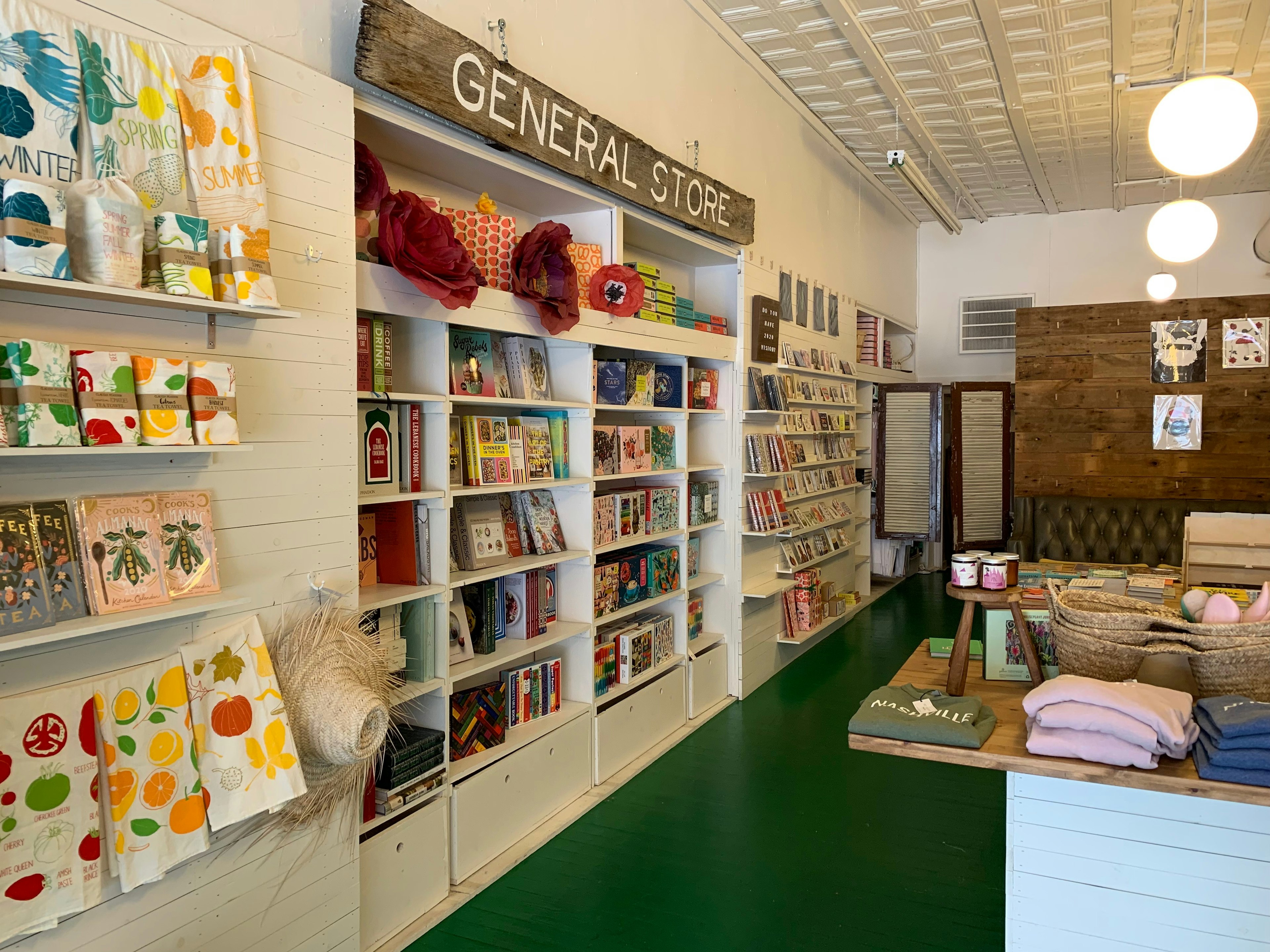 The interior of the Hey Rooster General Store shows colorful tea towels, books, t-shirts, paper flowers, candles, and more