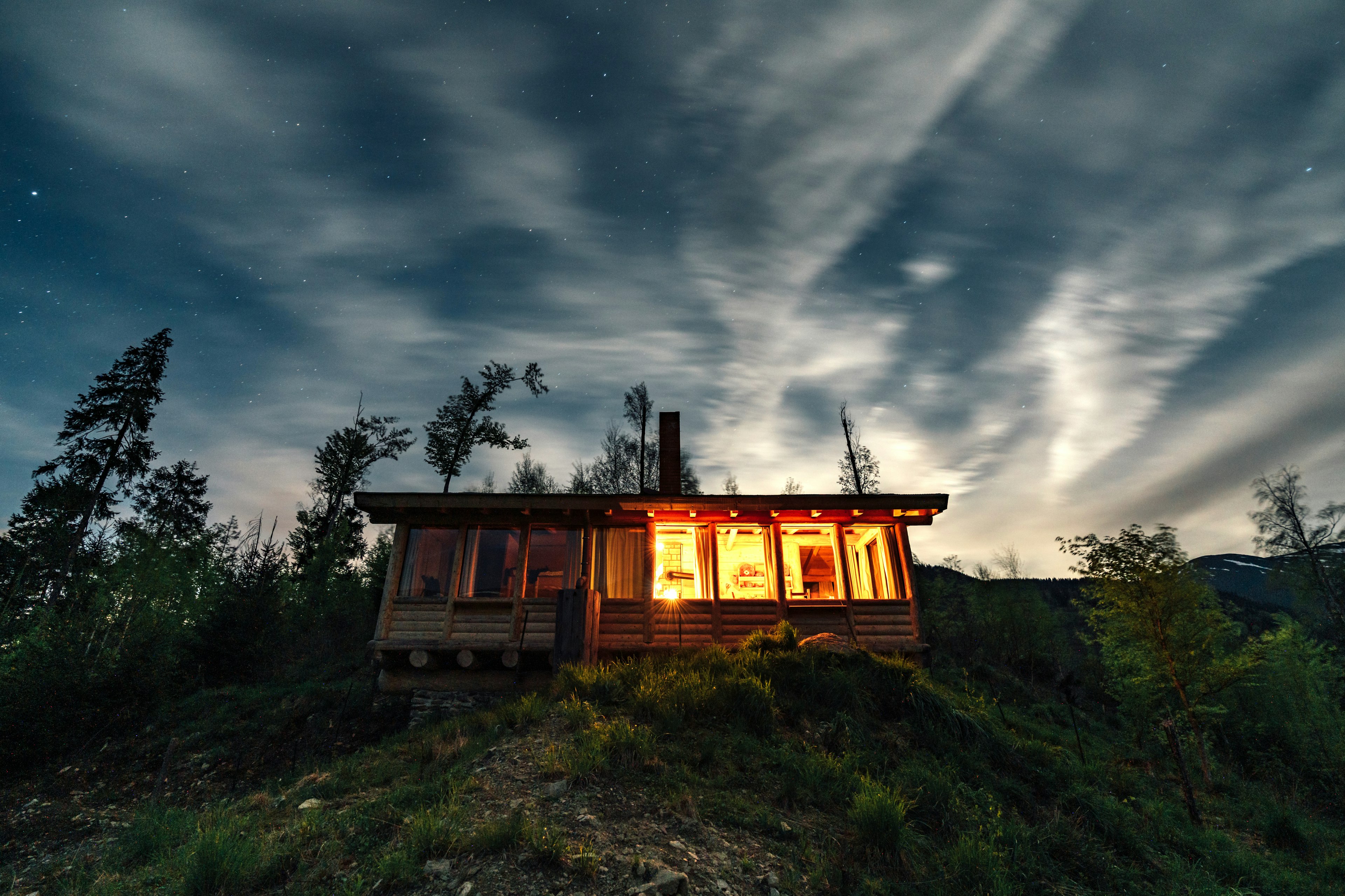 A modern looking wood cabin with sloping roof stands on a slope in the forest; half of it is lit up inside, giving it a golden glow in the impending darkness.