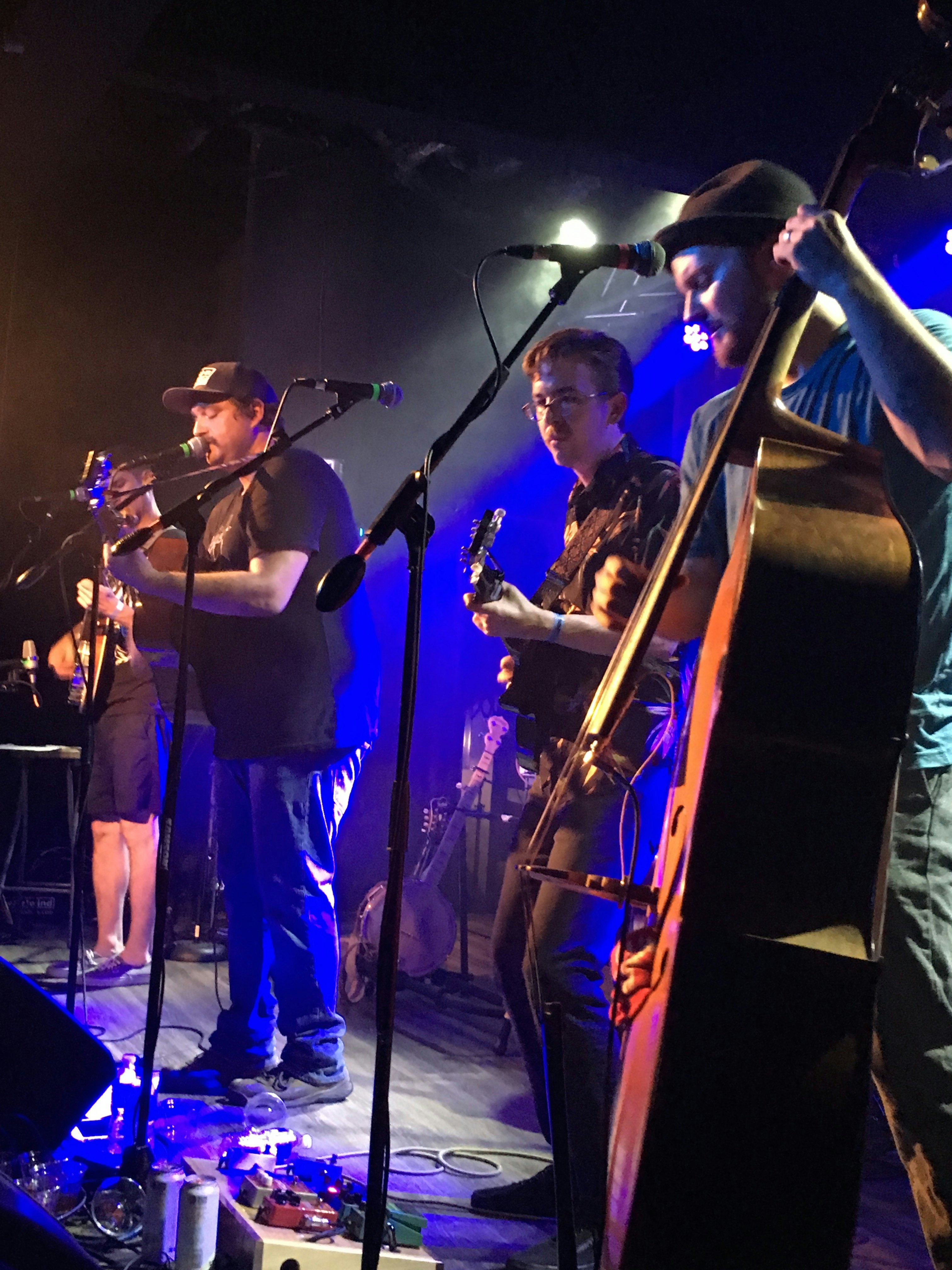 A group of musicians, each holding musical instruments perform on a stage at Hi-Fi. There man in the center wearing a black hat and shirt is singing into the microphone.