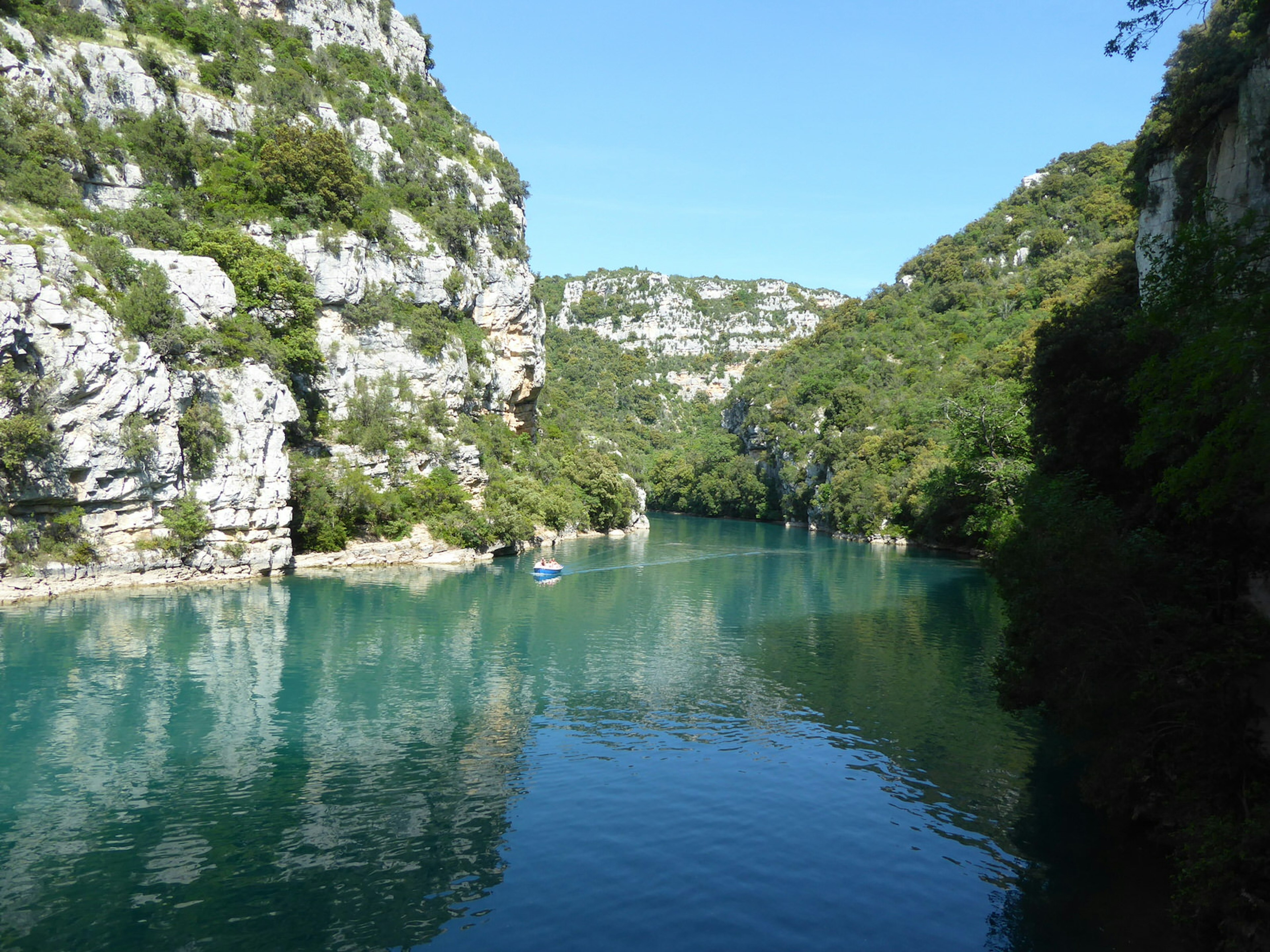 A close up of the gorge, the river is a bright green and there's a sailboat on it and the cliffs are high on either side with green bushes scattered throughout them.