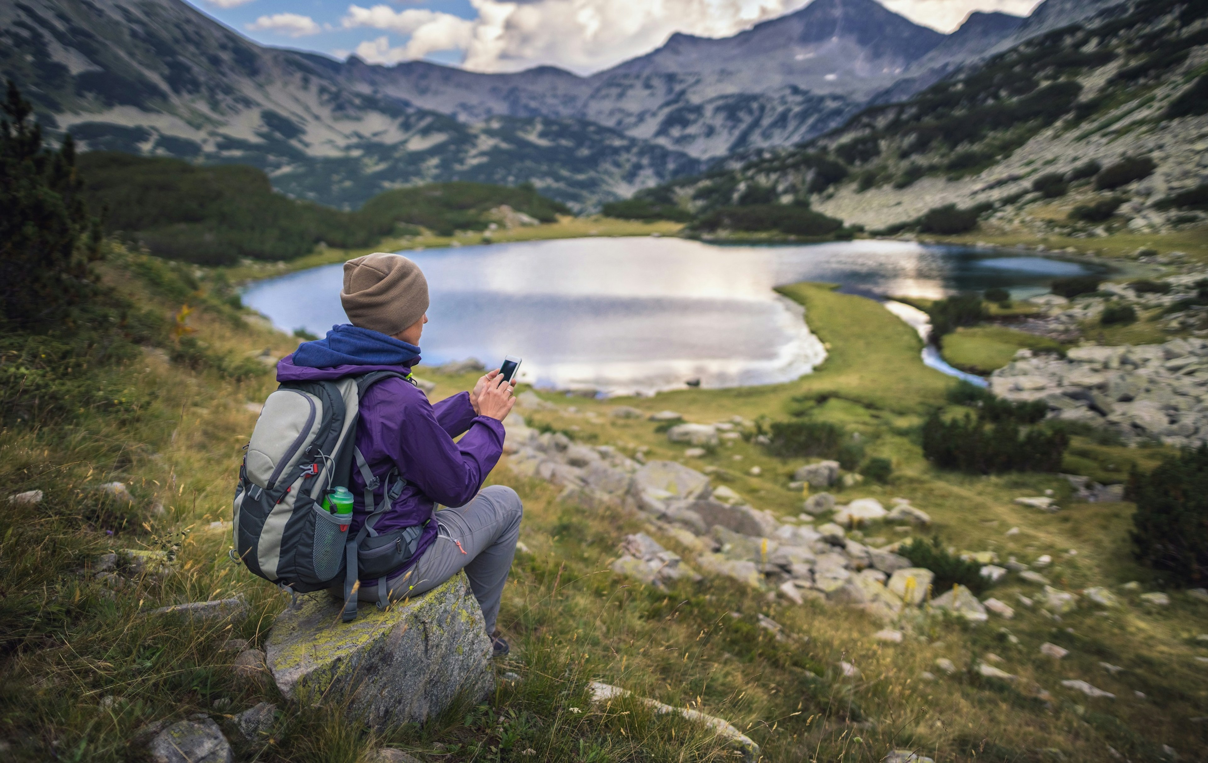 A woman hiker sits on a rock overlooking a lake, which is surrounded by circular ridge of rocky mountains; she's looking at her mobile phone.