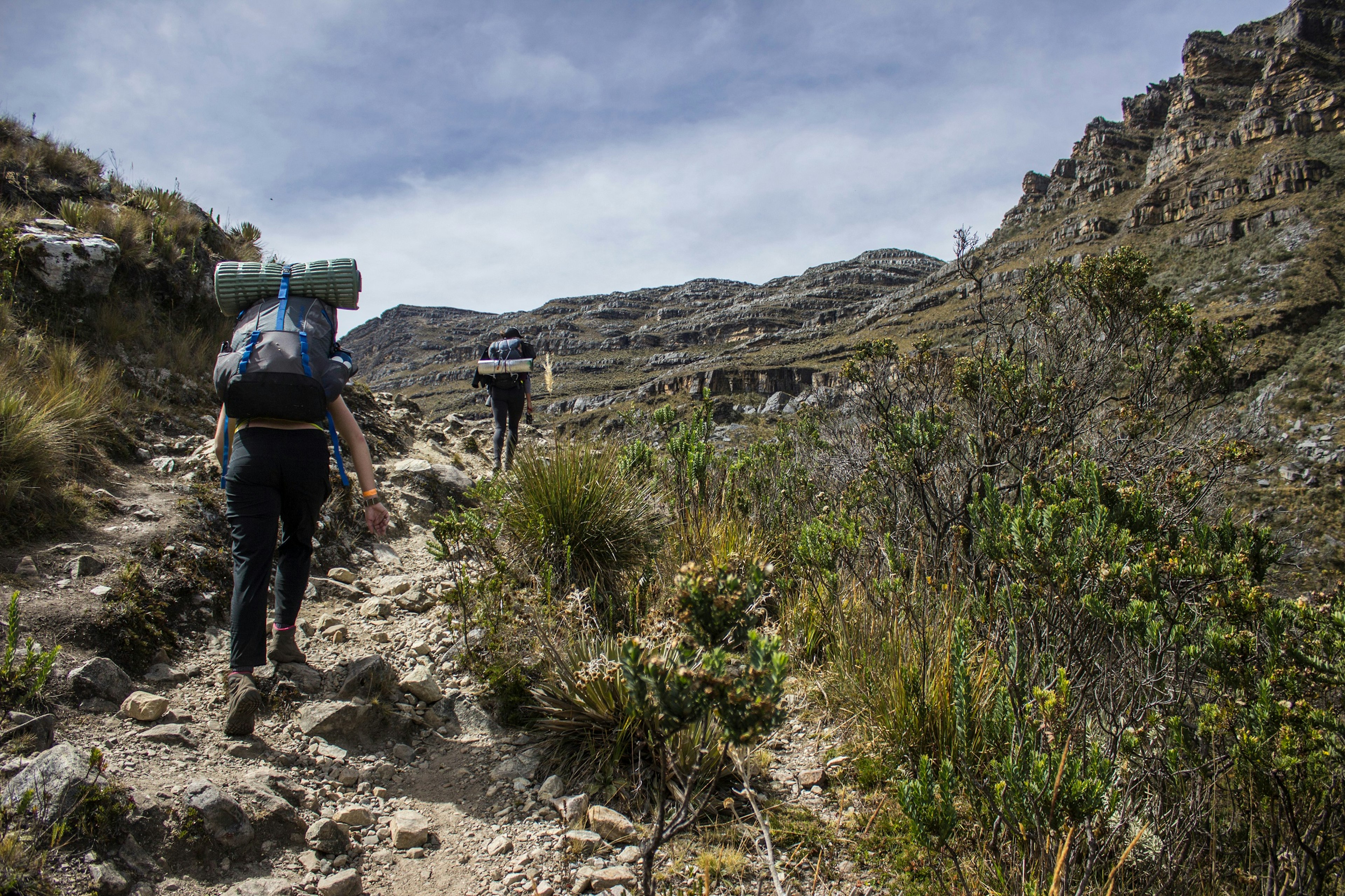 Two hikers, both wearing huge backpacks complete with sleeping mats, climb up a rocky trail within the mountains; the scene is quite barren, with no trees, just low shrubs and rock outcrops.