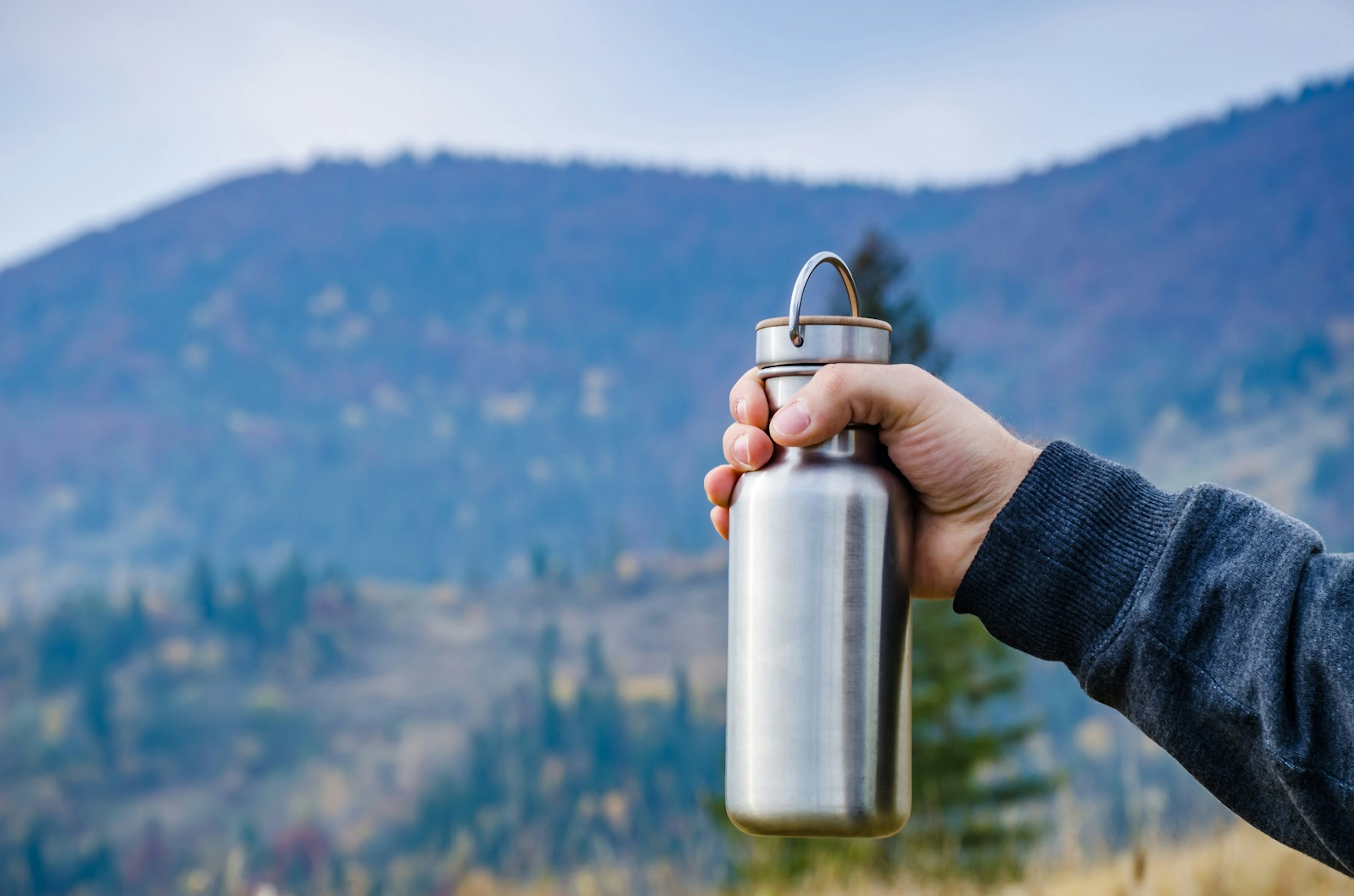 A hand holding a steel water bottle in front of rolling hills covered in coniferous trees.