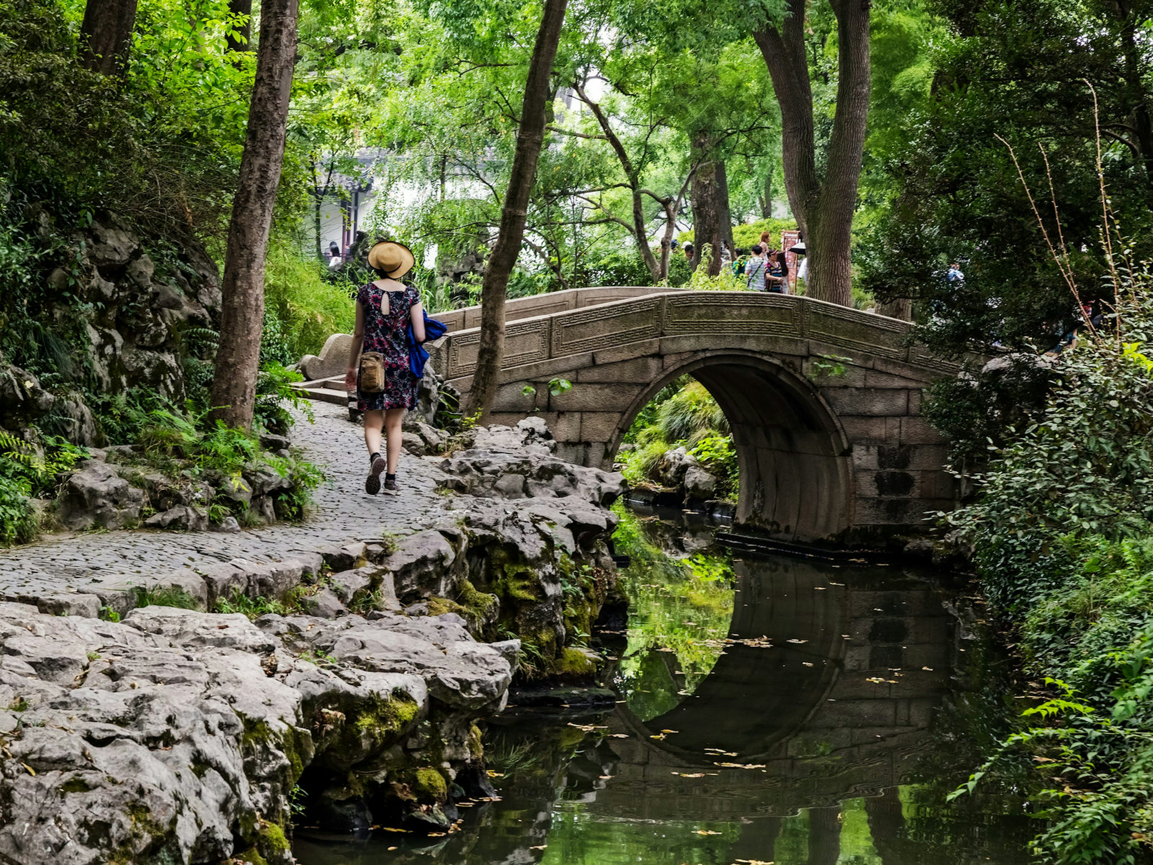 A tourist walks along a rocky forest path next to a pond with stone bridge. The serene paths of the Humble Administrator's Garden were restored in the 1950s © posztos / Shutterstock