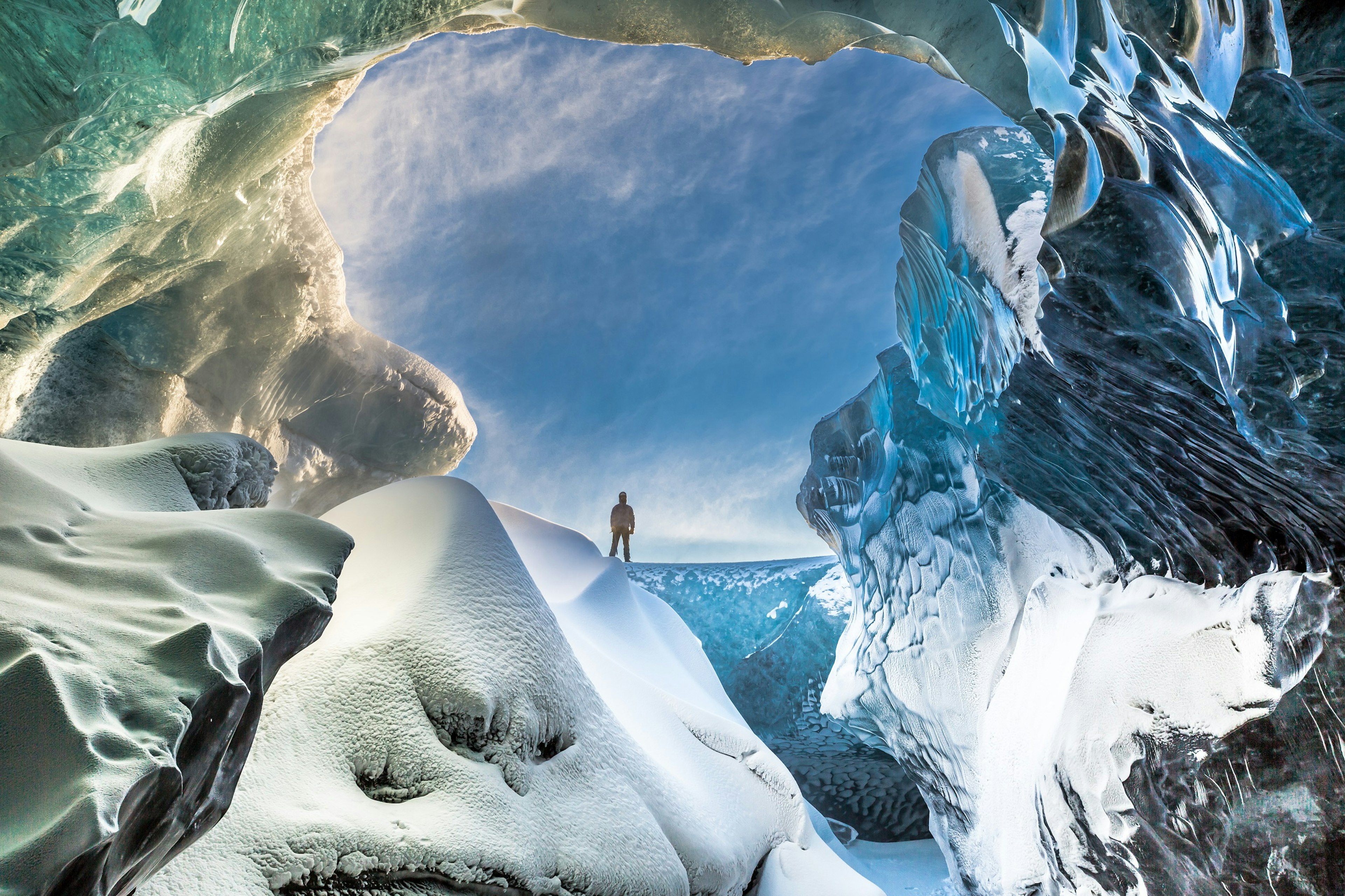 A shot from inside the Breidamerkurjokull ice cave in South East Iceland; it's looking out to a person standing on a slab of blue ice that looks like a crashing wave of water. The cave's walls glisten blue and green from the reflection of light through the ice.