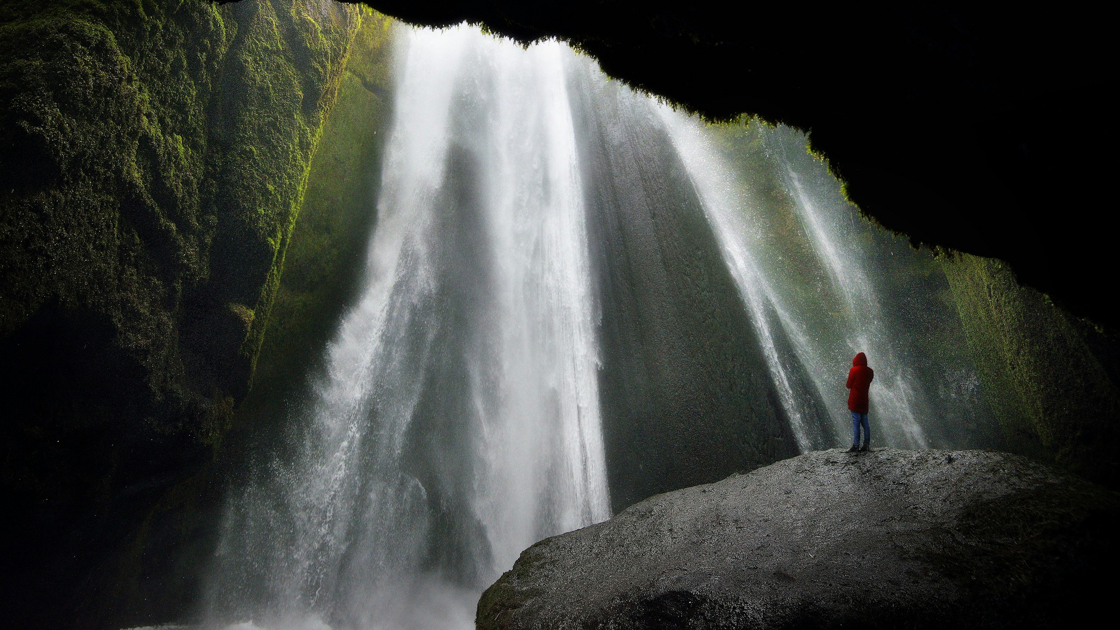 A lone person in a red jacket stands on a massive rock and stares up to a thundering waterfall crashing down in front of them; the walls of the cave are covered in vegetation.