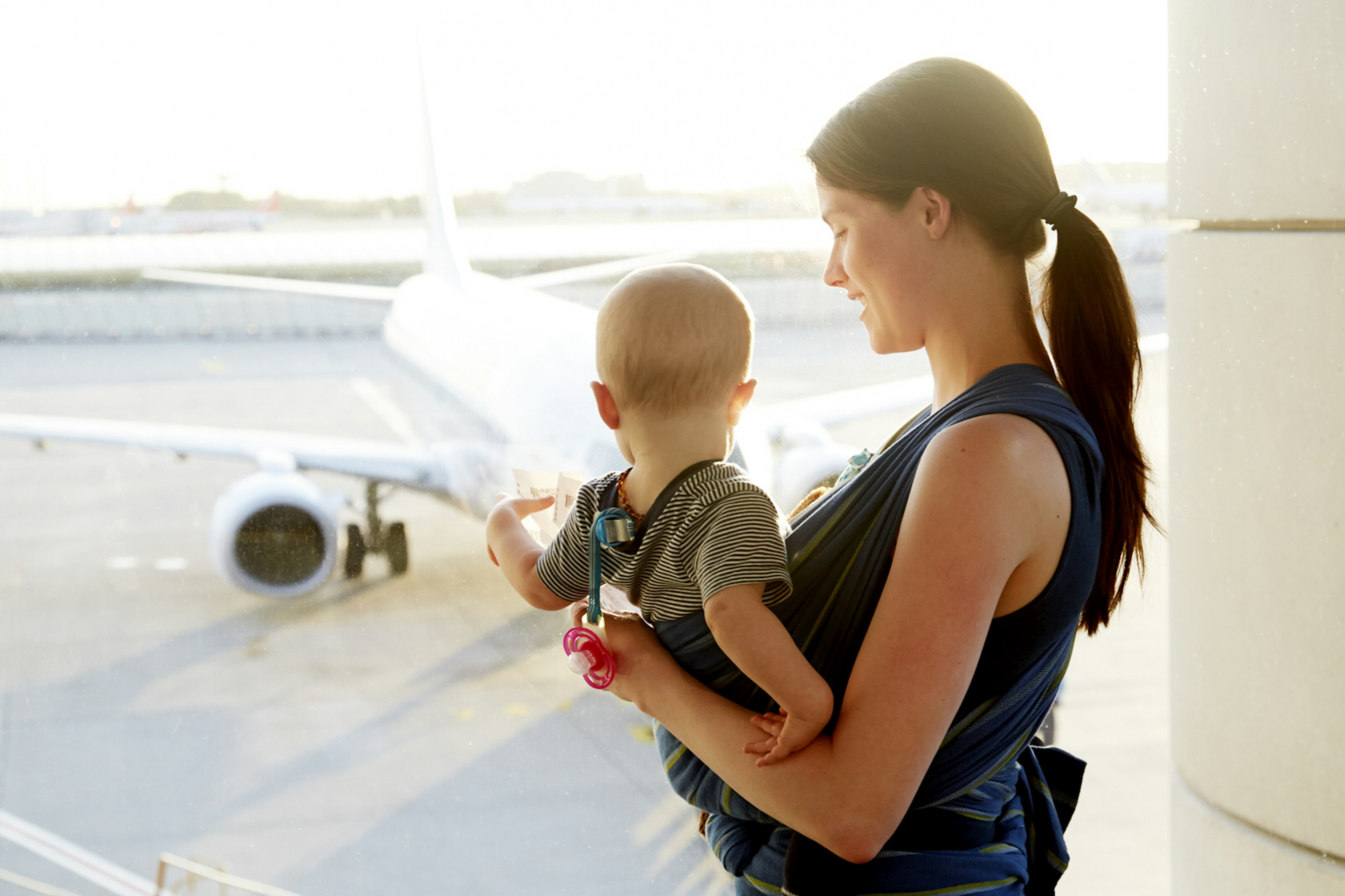 A mum looks lovingly down towards her young baby in a sling on her chest. The baby is looking away from the camera and out a window in the airport to a jet on the tarmac