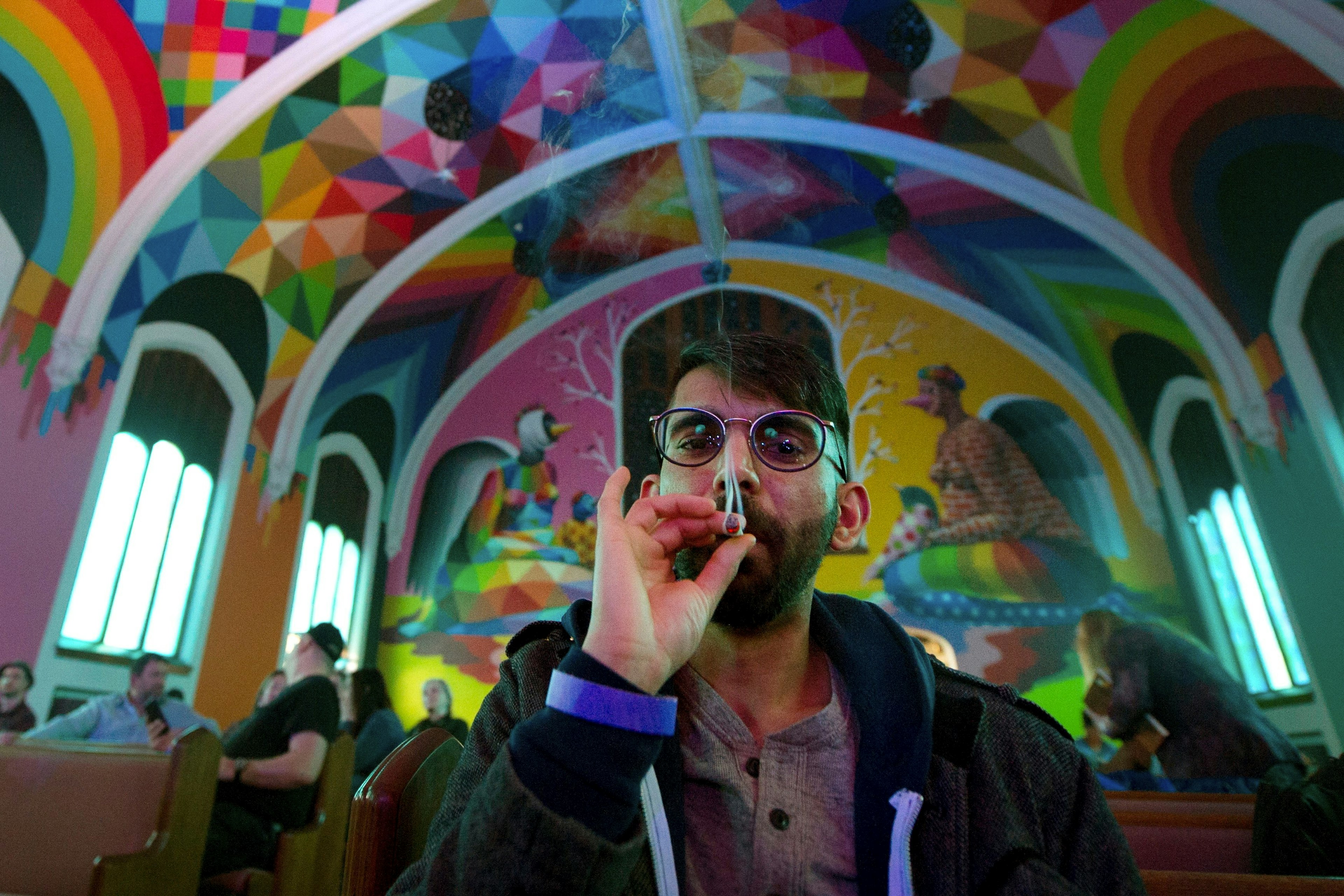 A man with glasses smokes a joint while gazing slightly down at the viewer. He is framed by the rainbow arched ceiling of the International Churhc of Cannabis. Behind him are other people sitting in pews for an Elevationist non denominational service