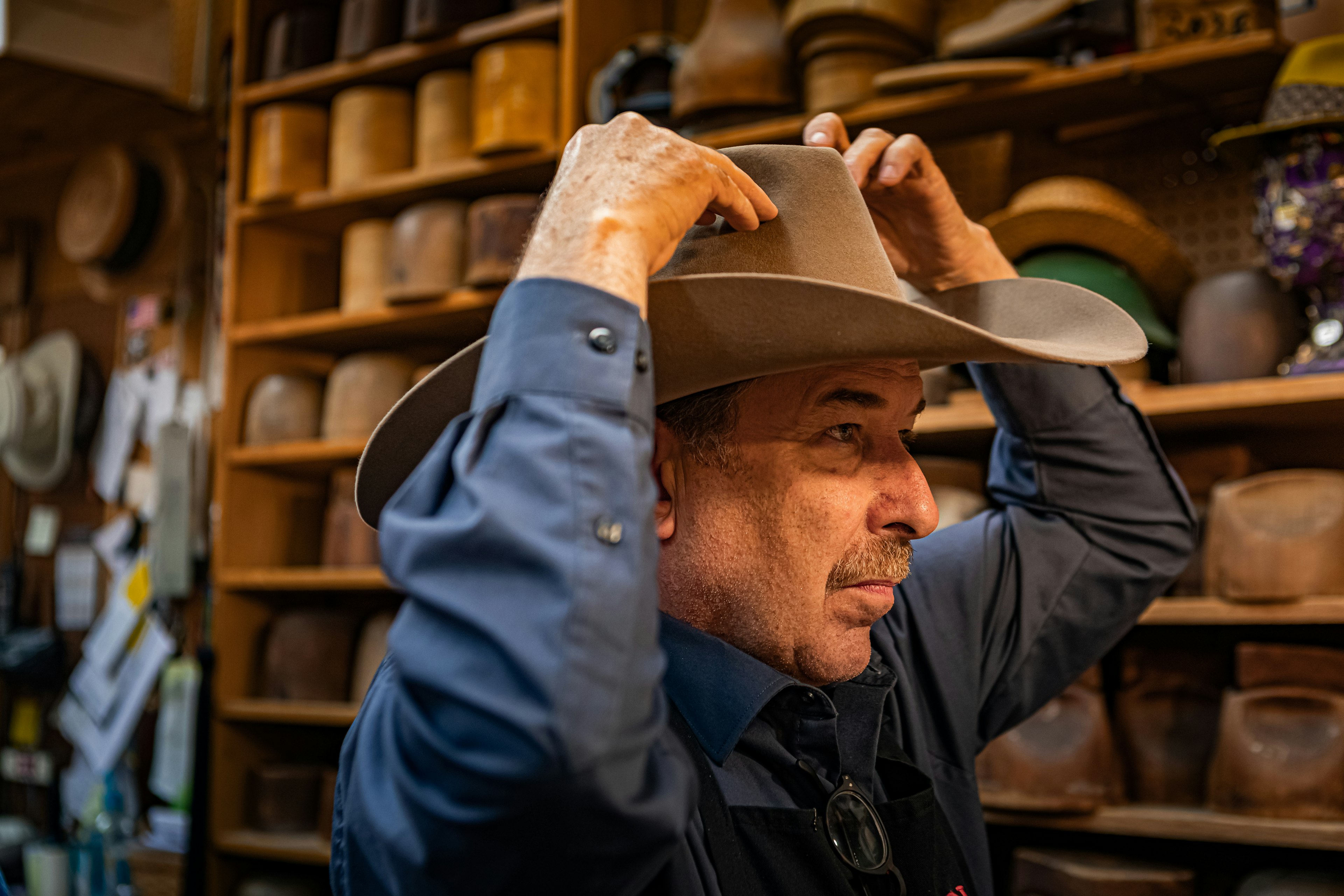 Jack Kellogg stands in profile to the viewer as he adjusts a large Stetson cowboy hat in a light brown color. In the background are many wooden hat molds on shelves