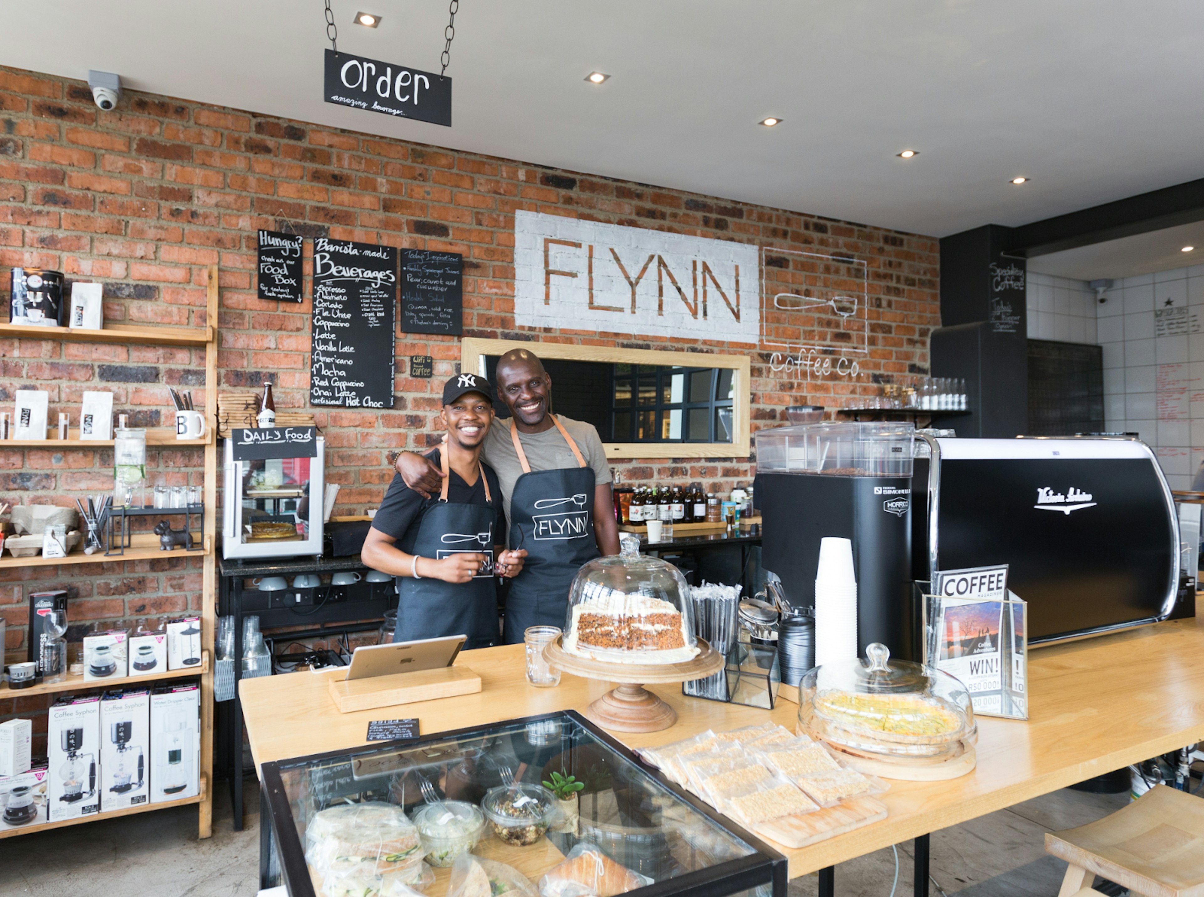 Two employees of Flynn Coffee stand smiling with arms around each others shoulders, with espresso machines behind and cakes in front. All backed by a retro brick wall.