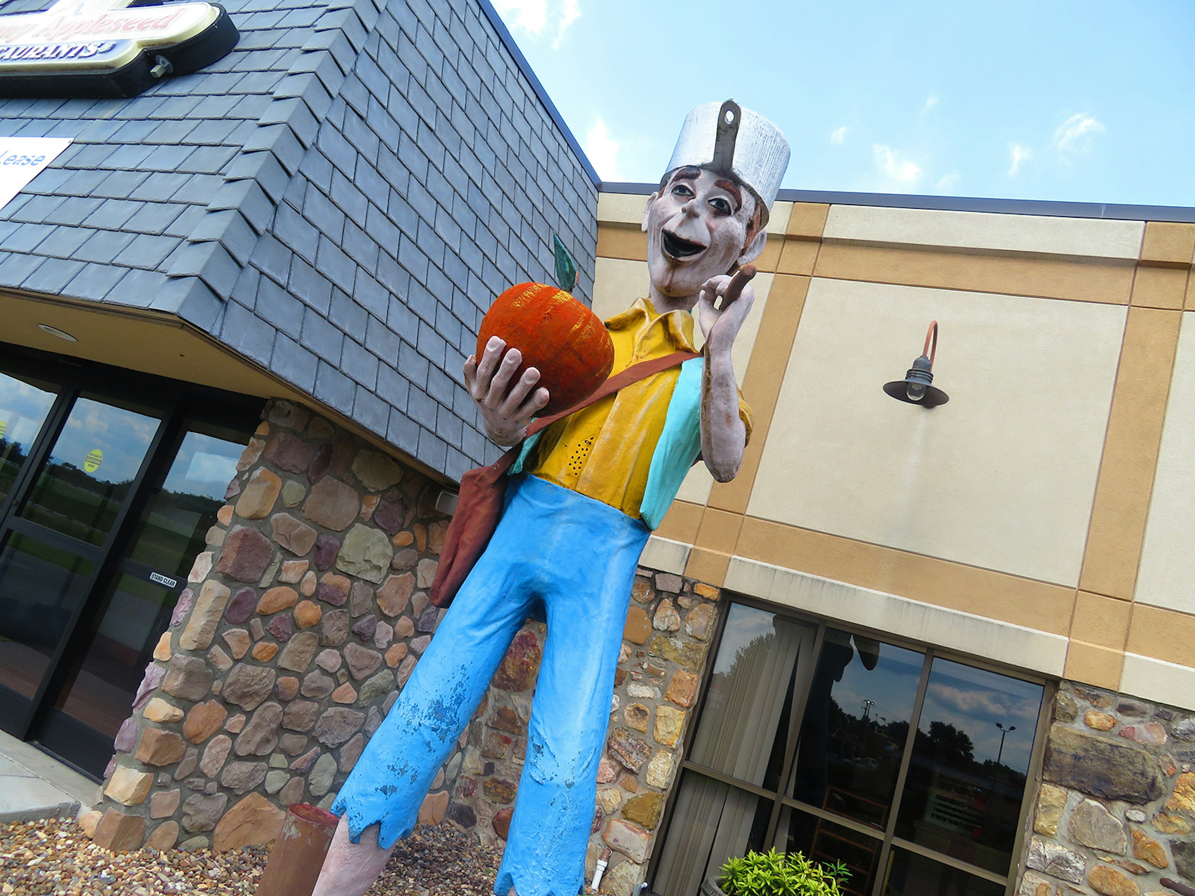 off-center photo of a statue of folk hero Johnny Appleseed, which stands 14ft tall. He's wearing blue pants, a yellow shirt and a frying pan hat – and he's carrying an apple.