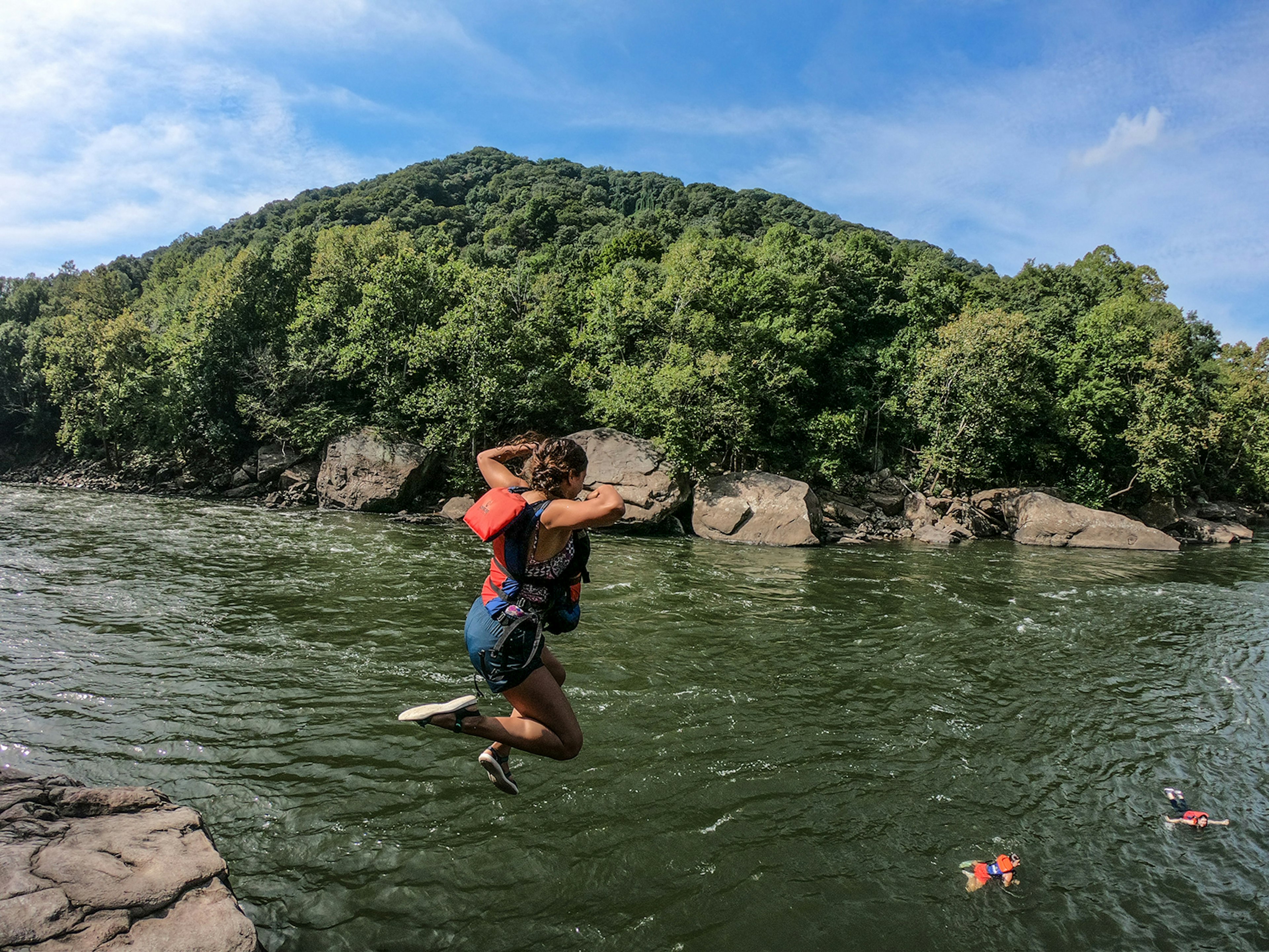 A woman wearing shorts and an orange life jacket jumps from a high sandstone rock into a river on a sunny day.