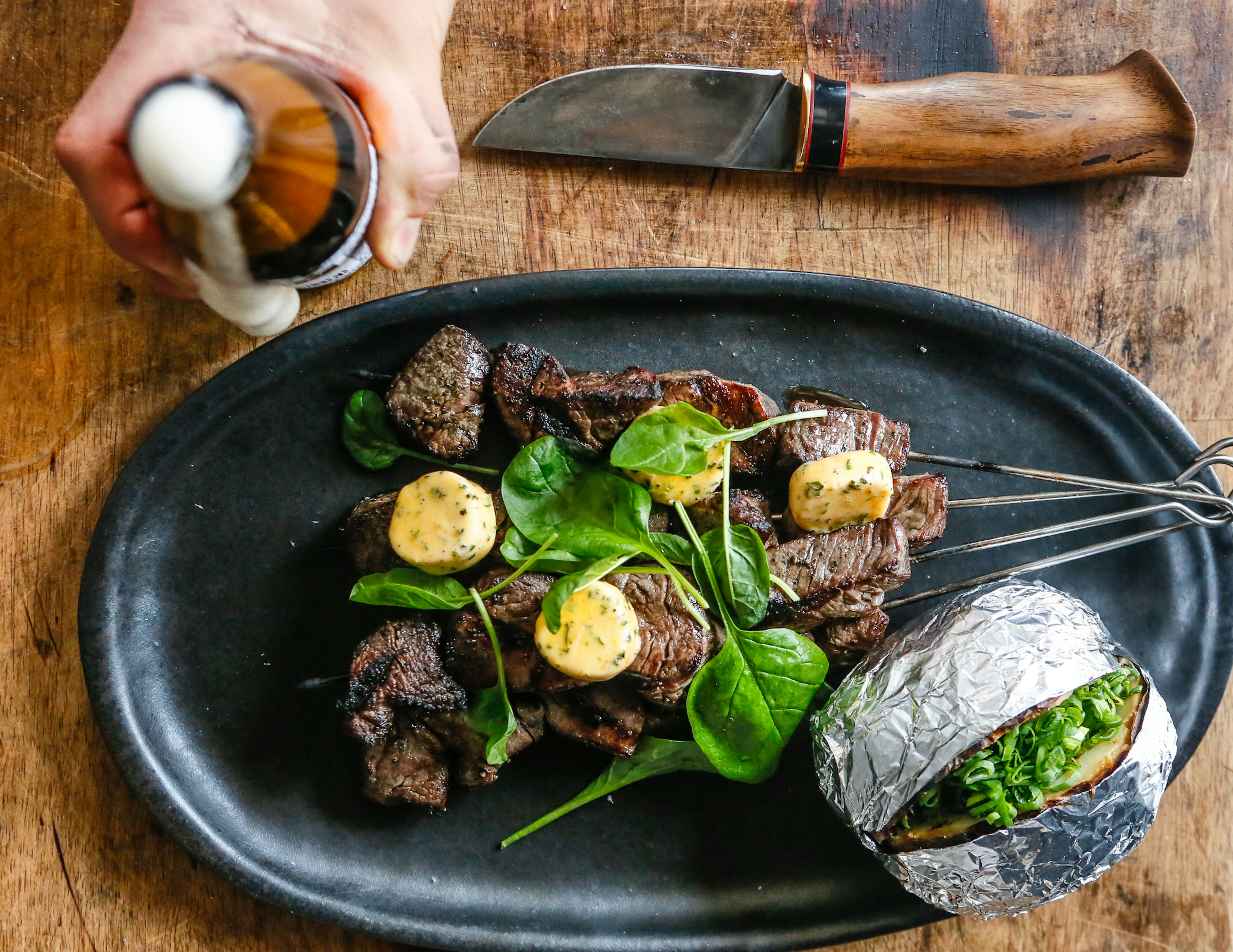 Looking down on a rustic meal of grilled meat skewers, and a jacket potato on a black oval plate at Kakolanruusu restaurant. A person is sitting at the table and holding a bottle of beer and a chunky knife sits beside the plate.