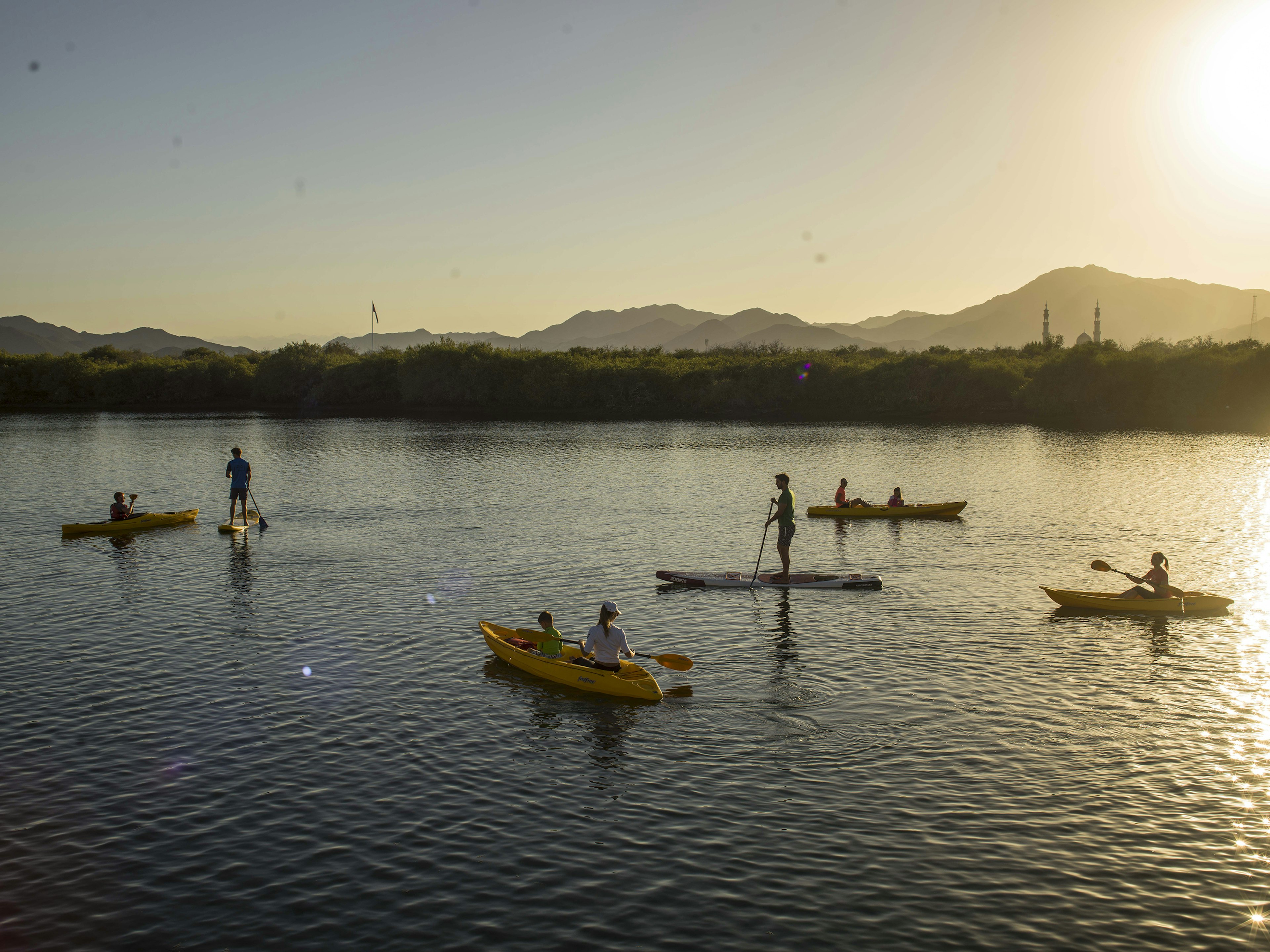 With the sun gleaming in the top right corner, eight people (two on stand up paddle boards, six in kayaks) play on the water of Kalba; in the distance is the shrub-covered shore.