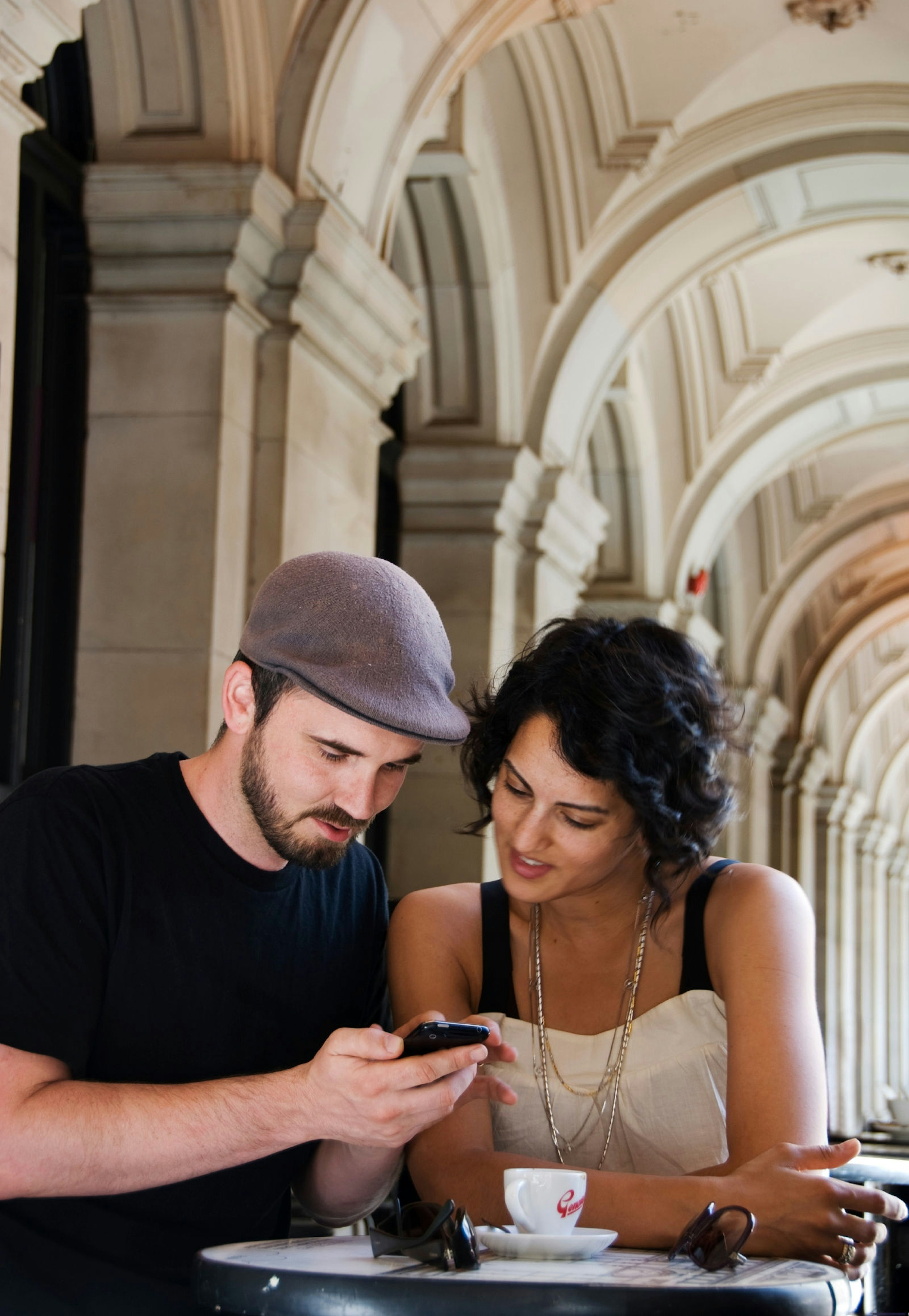 A young man wearing a black tshirt and grey hat plays with his phone while a young woman with black hair and a cream top looks at the screen. They are standing in a cafe with a coffee in front of them, leaning on a table.