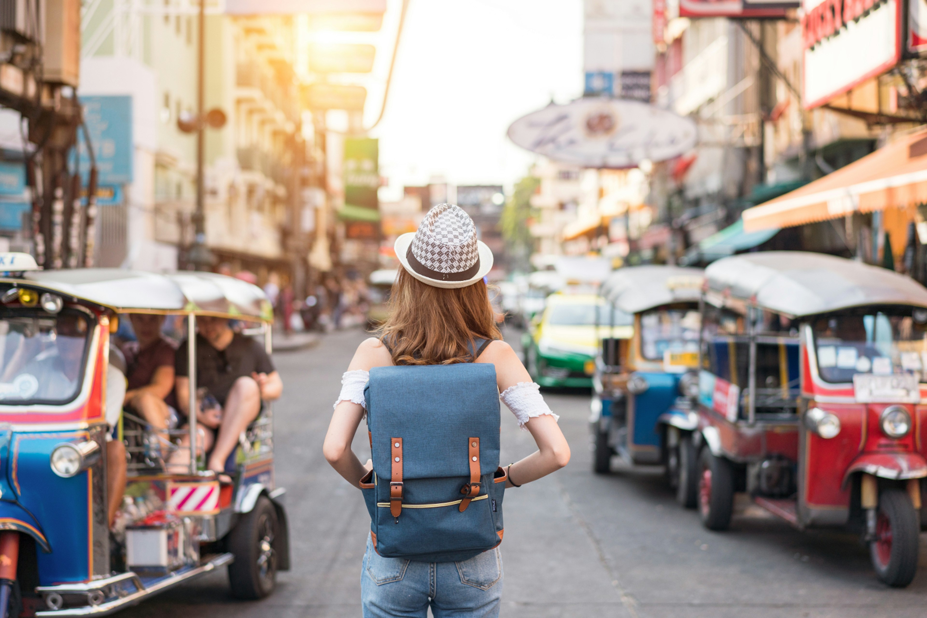 The back of a young woman walking along the pedestrian street in the evening in Bangkok, Thailand; travelers, tourists and tuktuks line the street.