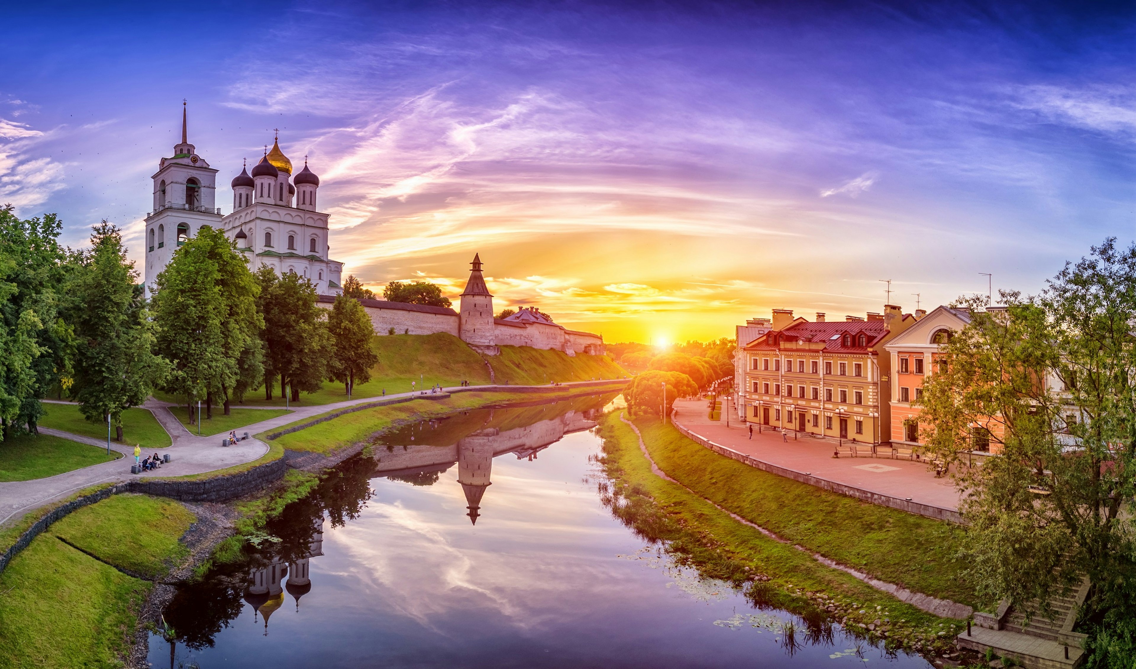 A river with a row of pastel-coloured buildings on the right bank, and fortress walls on the left bank. Within the fortress is a church with five domes, the central dome is gold