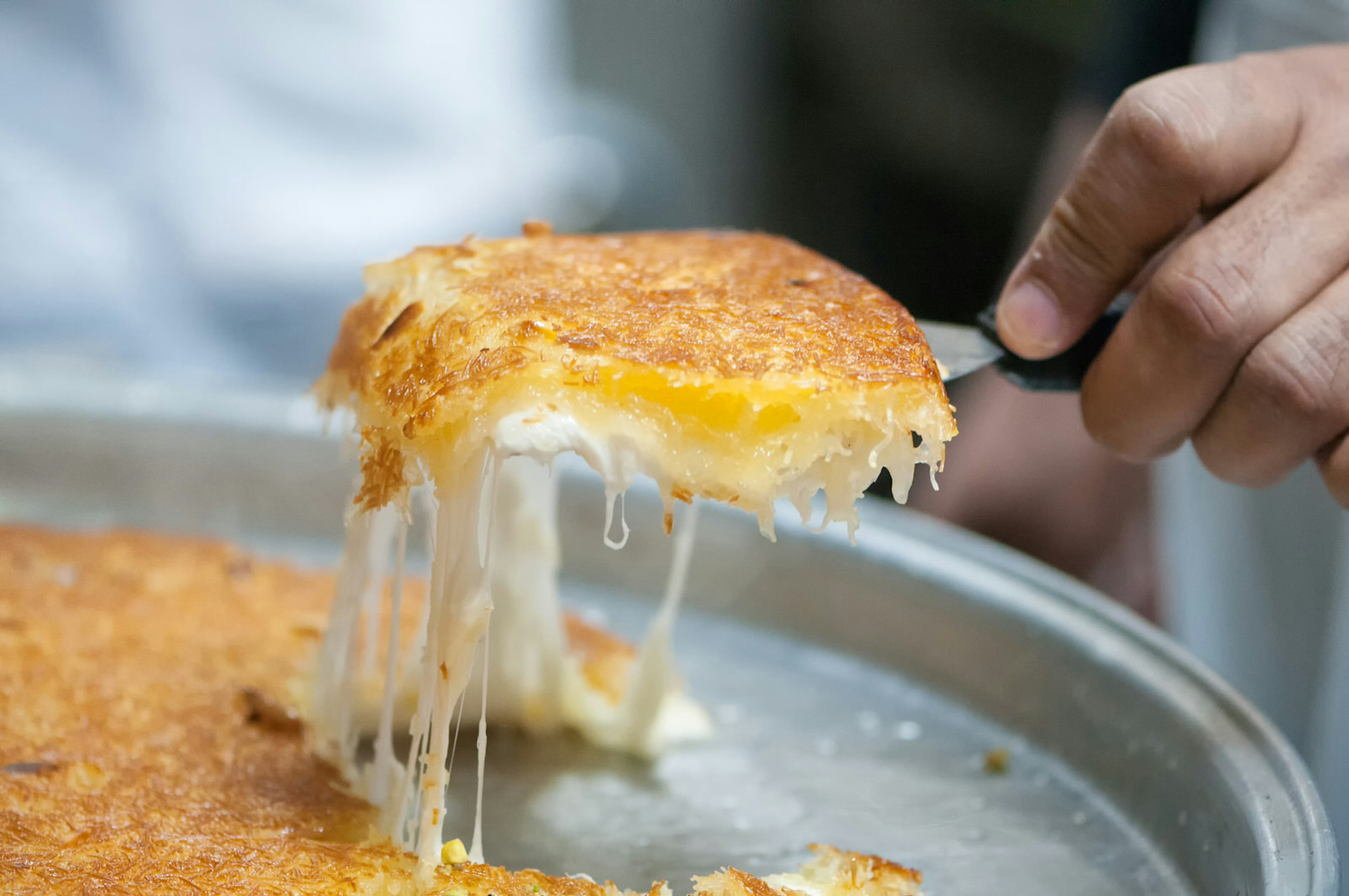 Close-up of a baker’s hand pulling freshly baked kunafeh pastry © PamelaJoeMcFarlane / Getty Images