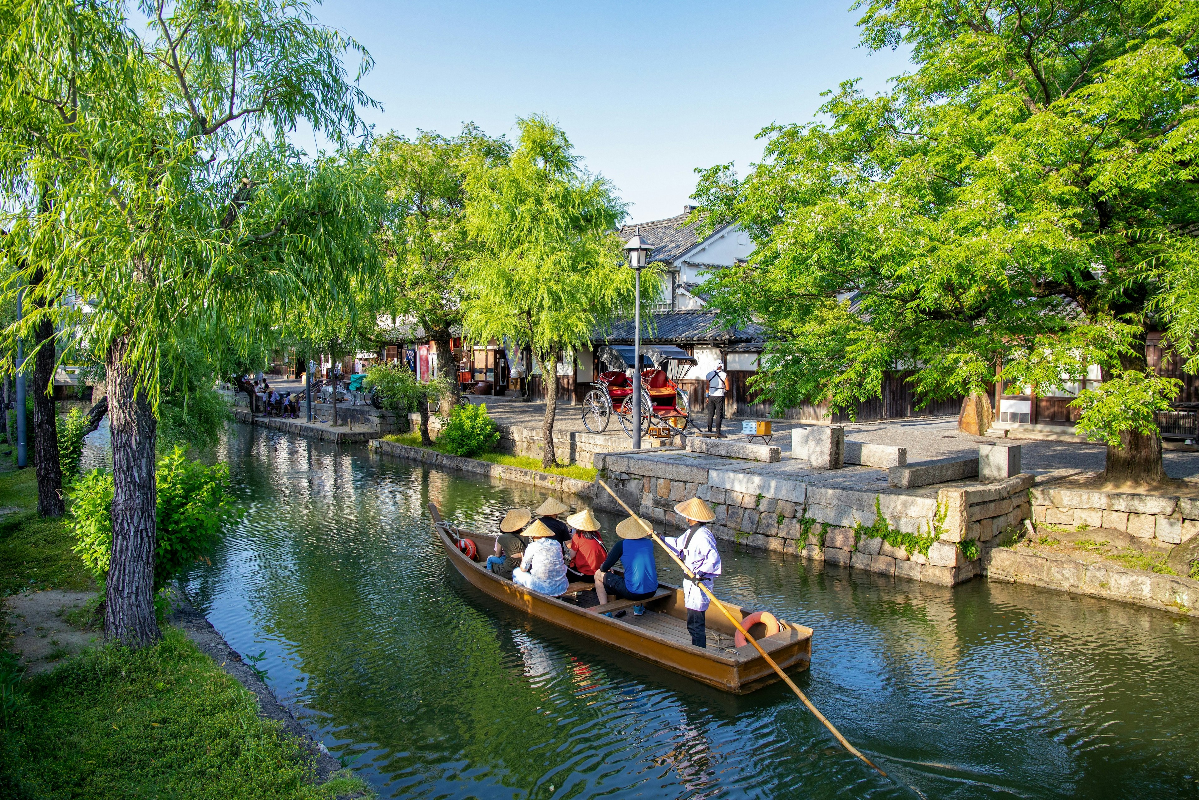 A man in a kimono rows a boat of passengers, all wearing conical rice hats, along a narrow river in the town of Kurashiki. The river is flanked by green trees.