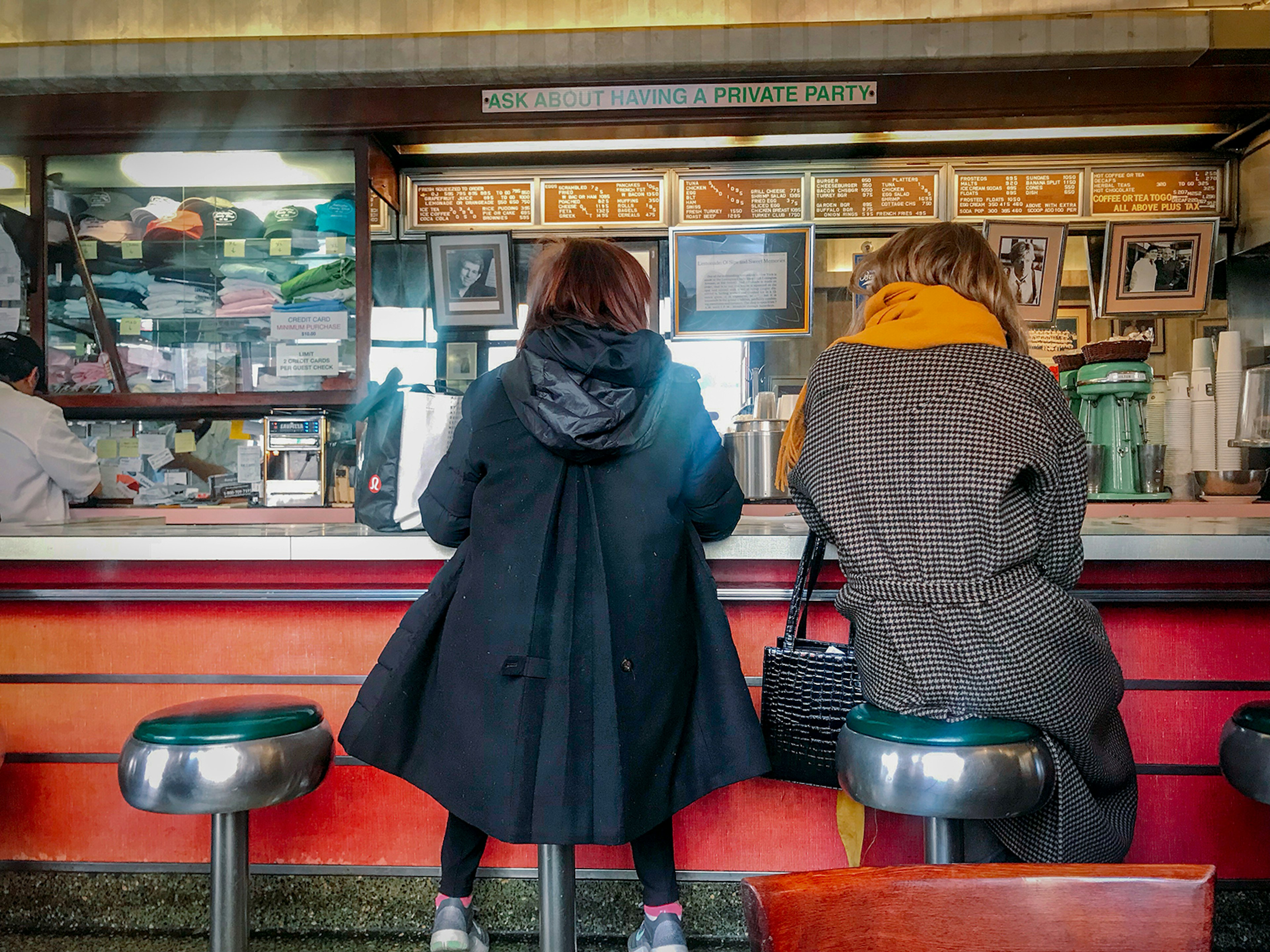 Two women wearing coats sit on vinyl-covered stools at a diner counter in New York City