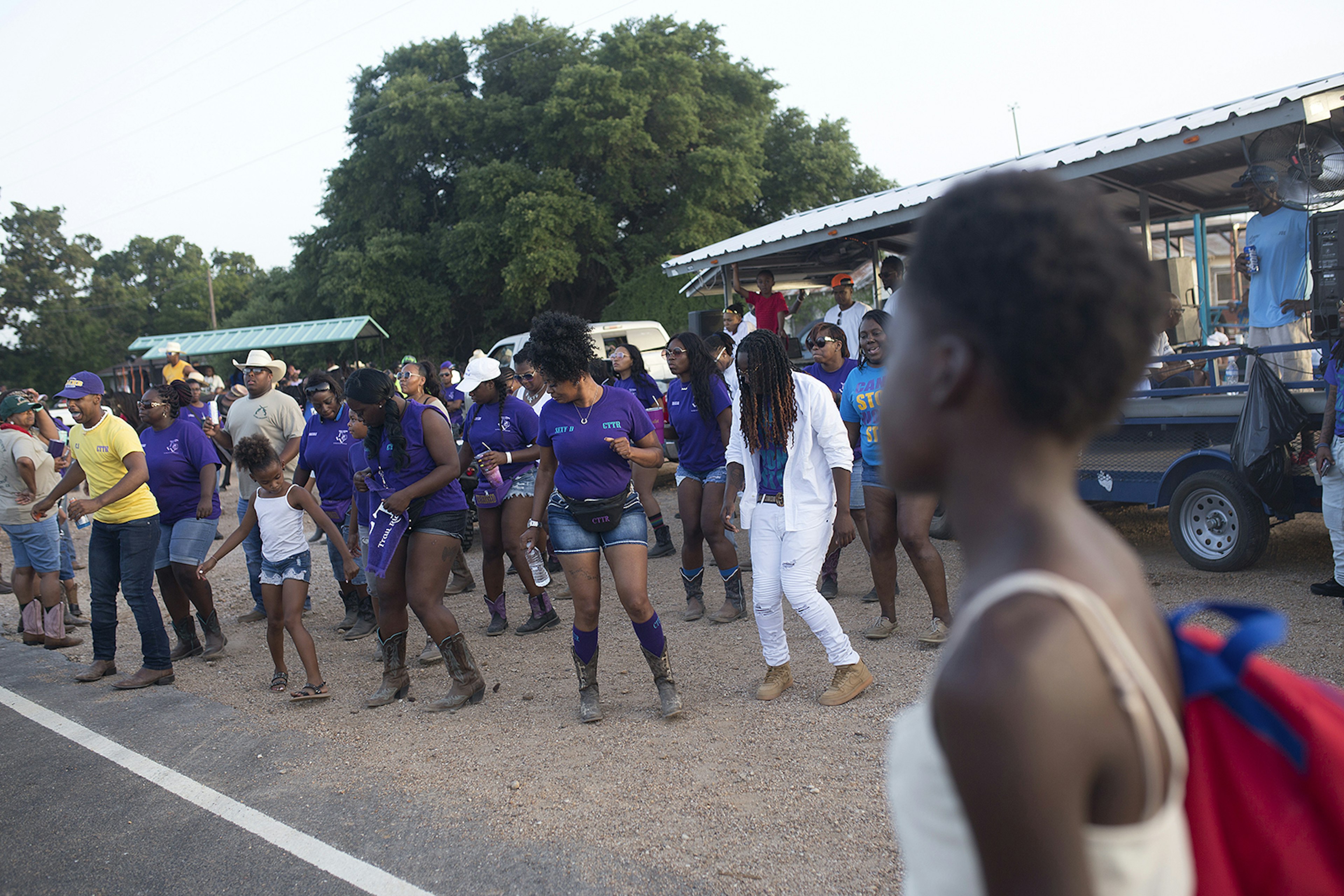 group of people dancing on a dirt shoulder next to a trailer fitted with speakers