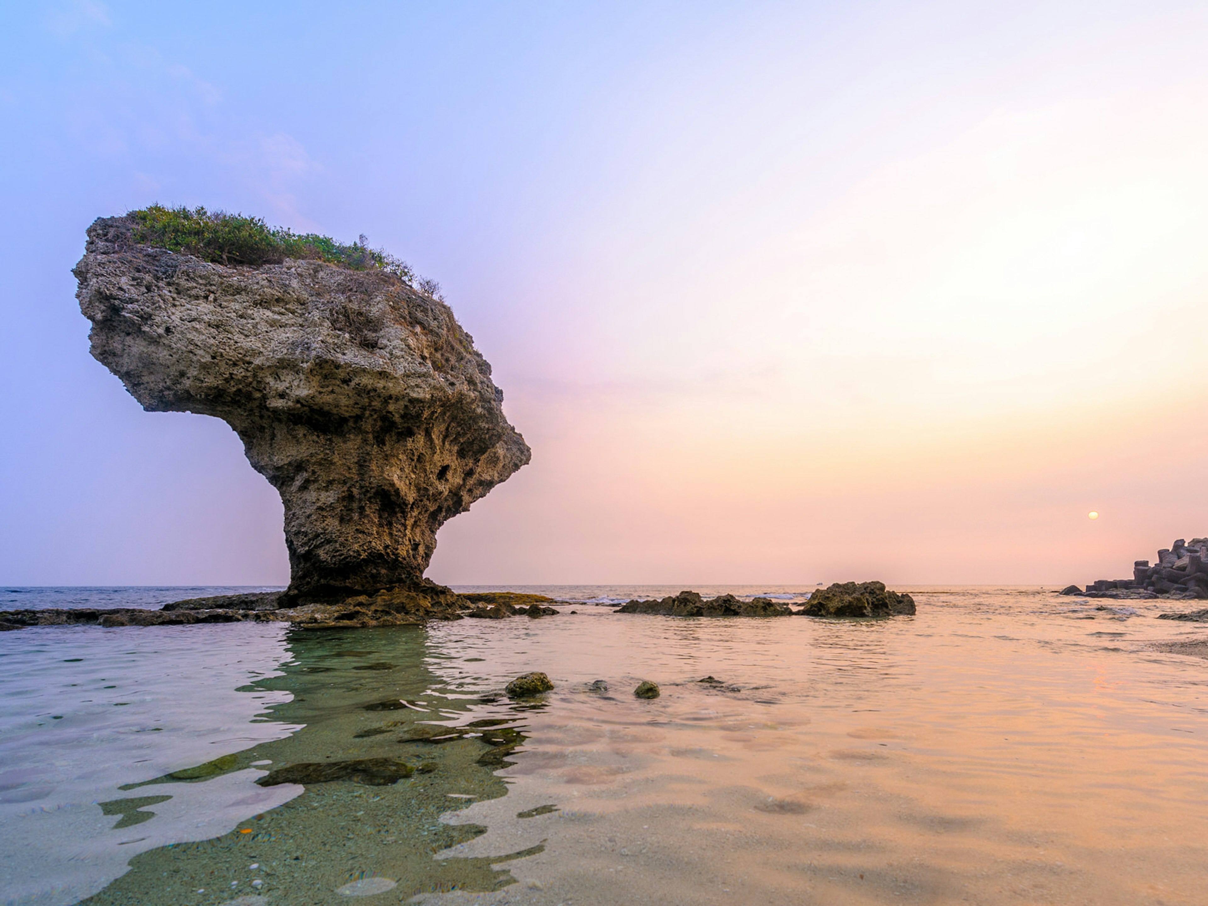 A large coral rock formation rises out of clear sea waters with pink and purple sunset and moon behind. Vase Rock is Little Liuchiu Island's star landmark ? Richie Chan / Shutterstock