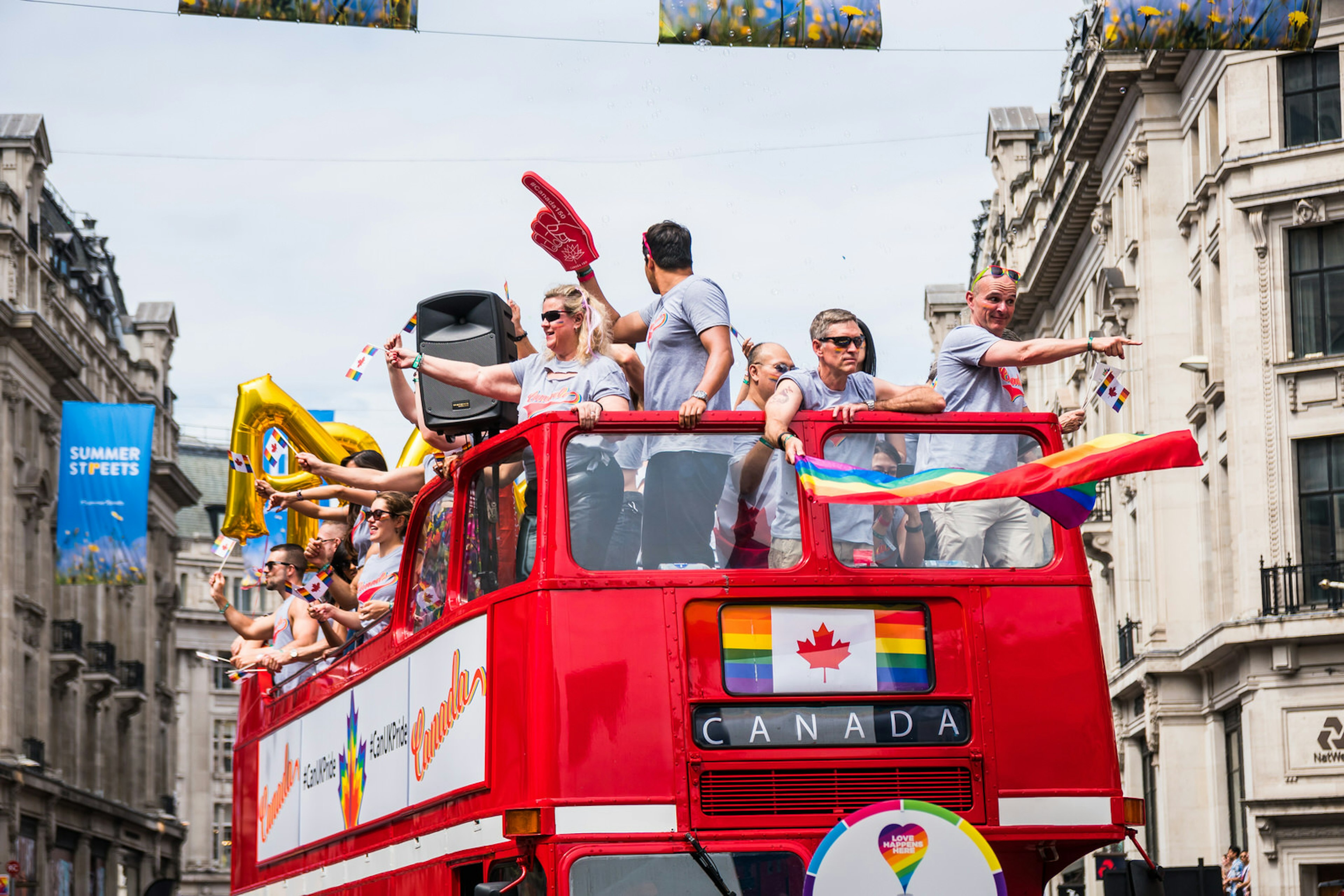 People on a topless double decker red London bus on the Pride in London parade on Regent Street; the bus' destination reads CANADA and there is a Canadian flag above it, with the normally red stripes replaced by rainbow colours