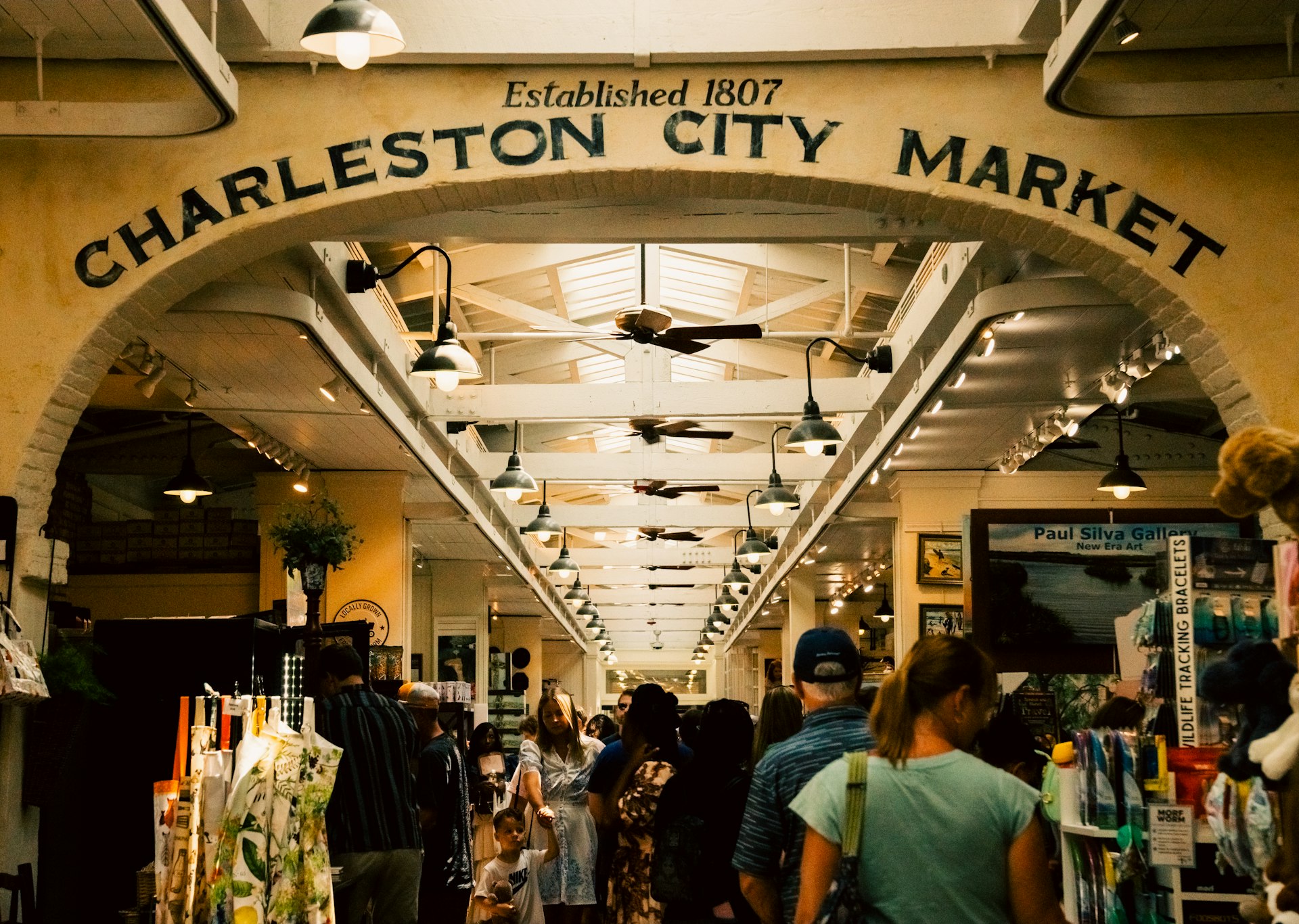 An indoor market with people shopping. Above, on an arch, reads a sign saying "Established 1807 Charleston City Market"