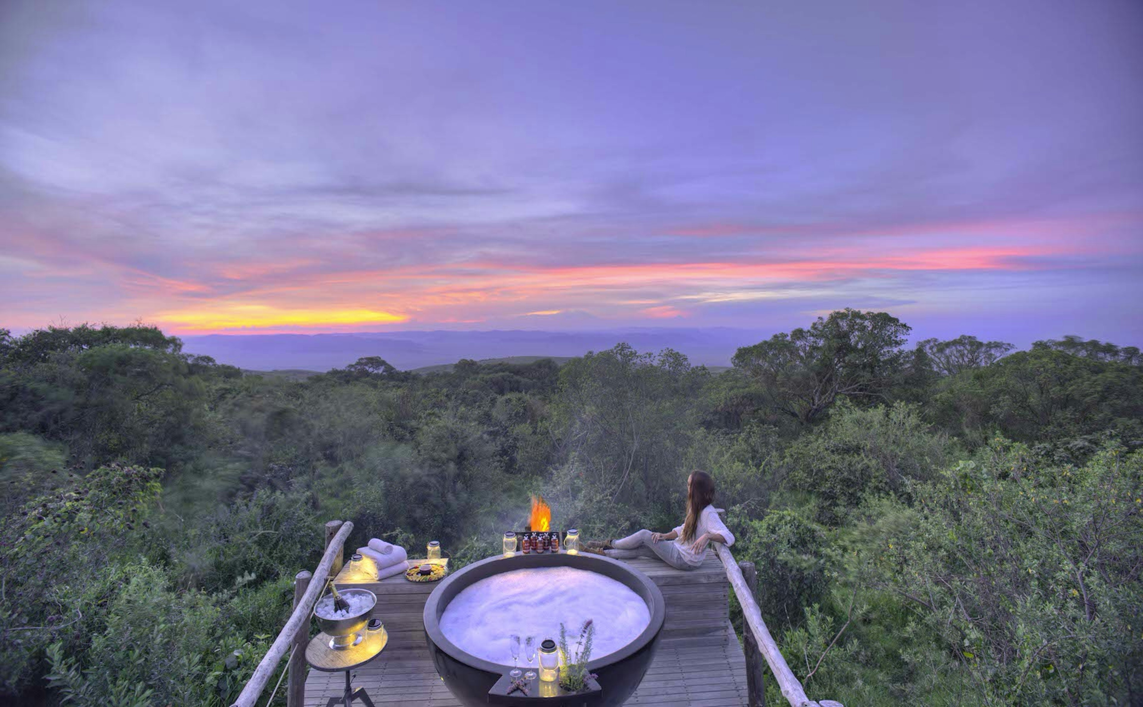 A woman reclines on a wooden deck and looks out on the wilderness from a wooden deck at sundown. The deck has a hot tub, candles and small tables.