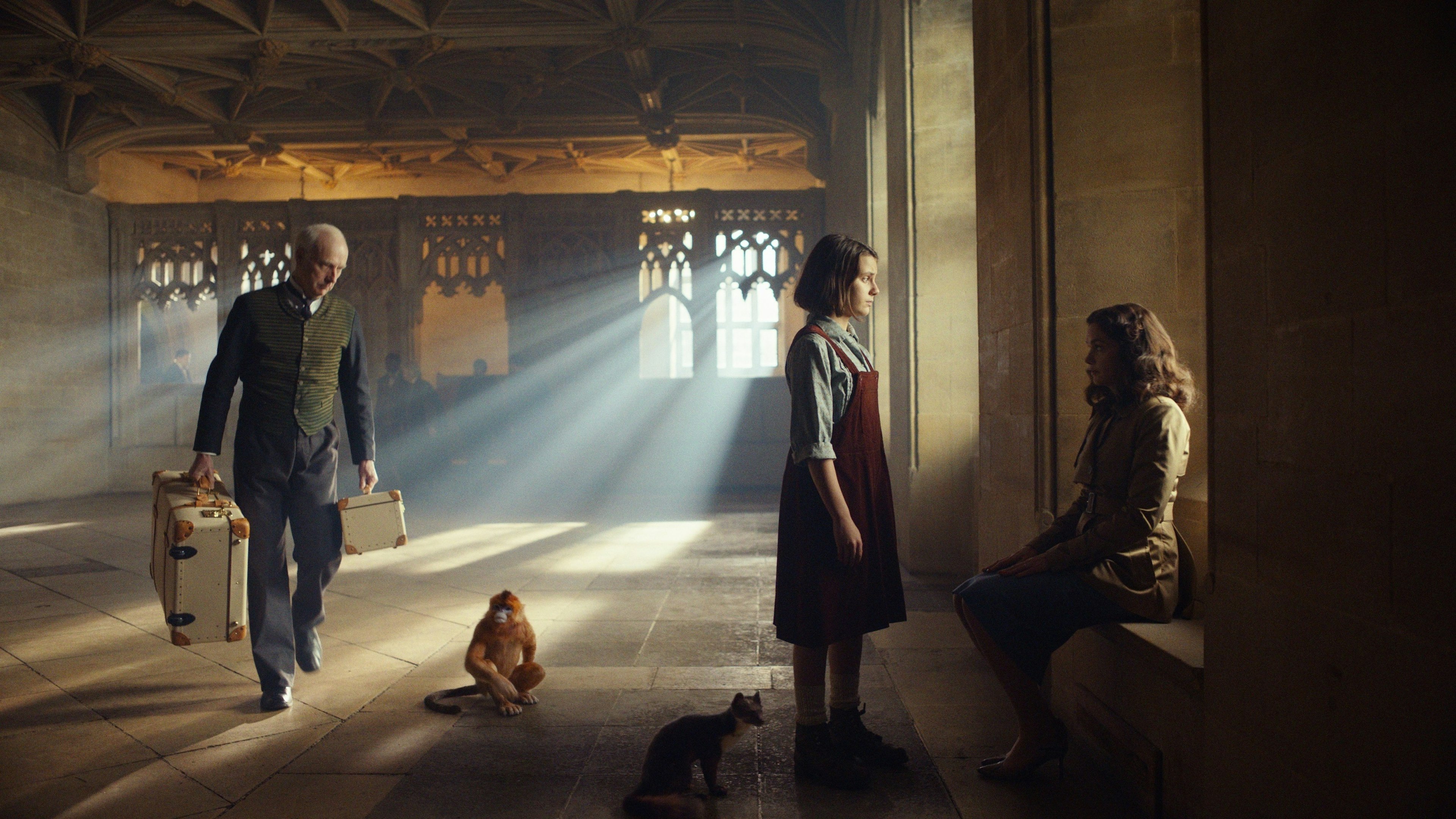 Lyra looks out a window while Mrs Coulter sits on the window. A servant is carrying bags and a golden monkey and a ferret are on the ground.