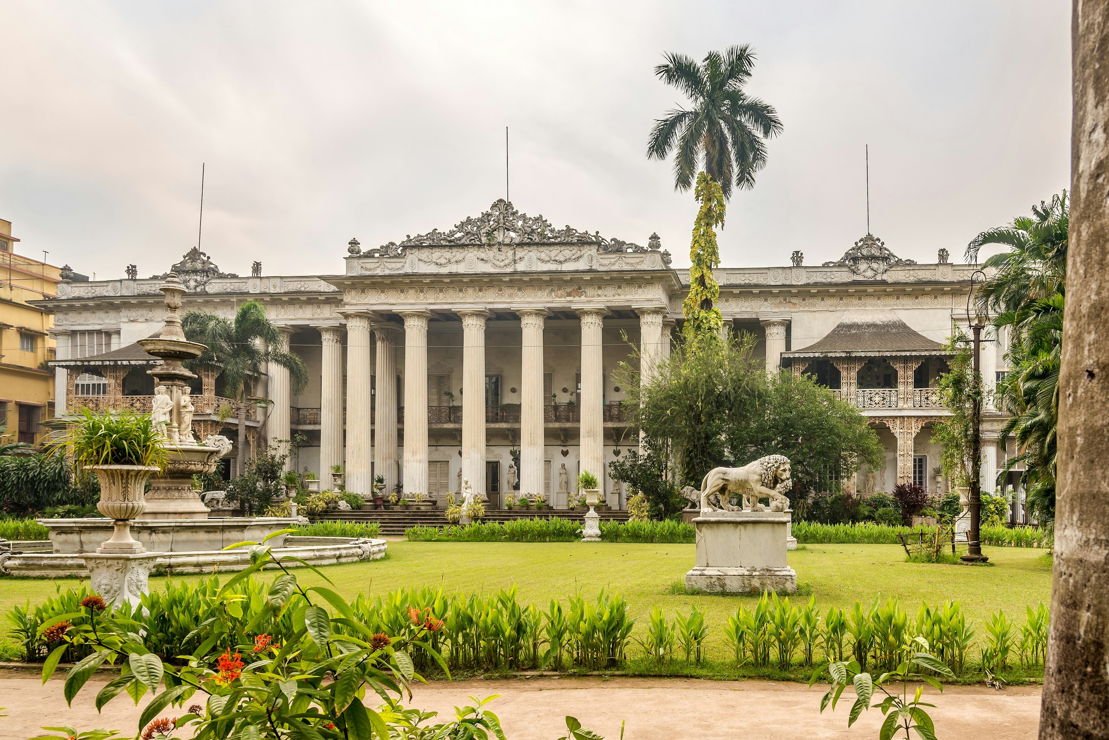 The exterior of Kolkata's Marble Palace as seen from the front. The palace is grand, with white columns and fountains in the green gardens.