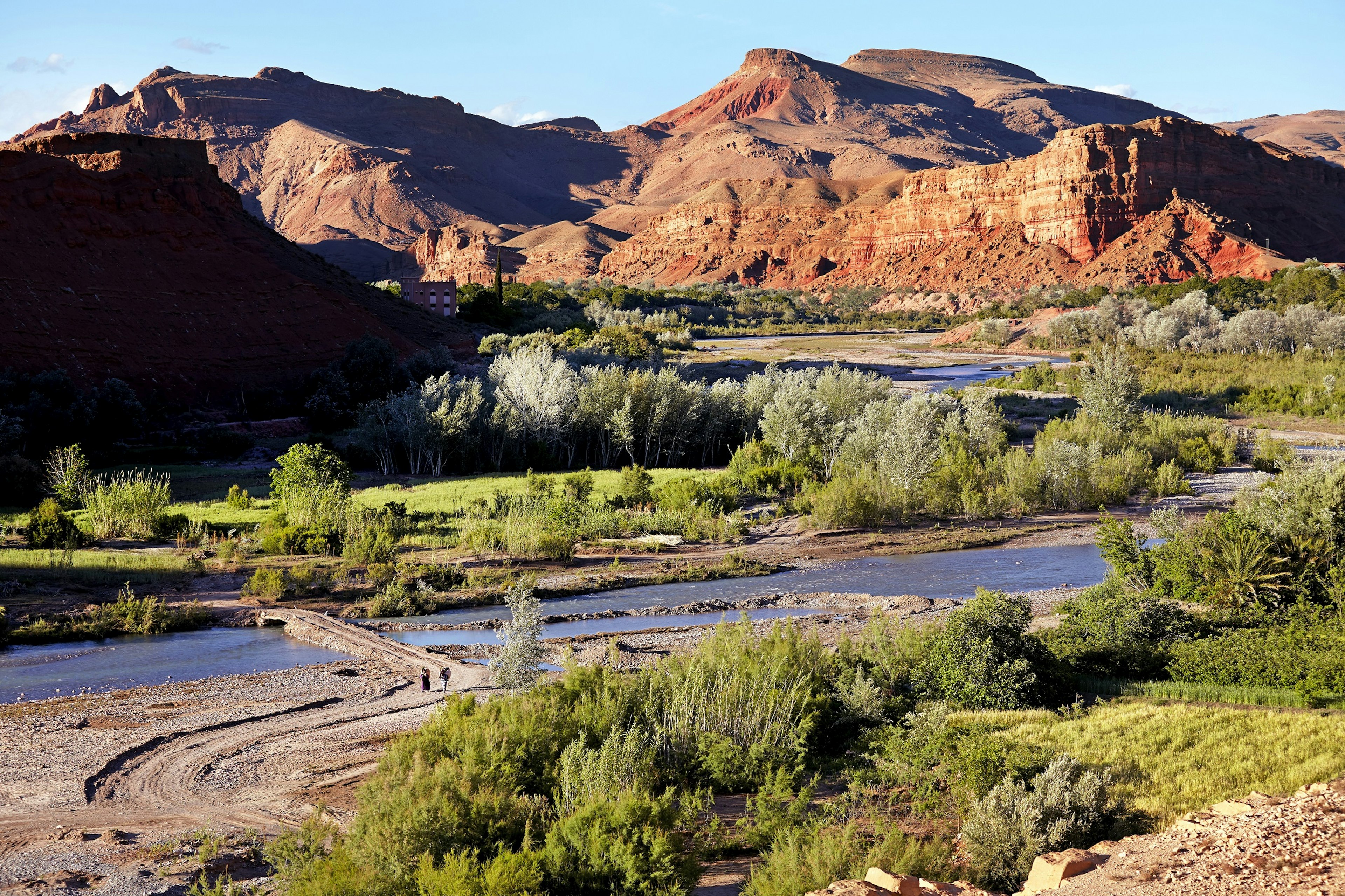 Two people hike along a dirt track that crosses a narrow section of river within the M'Goun Valley; trees and grass cover the lower valley, which abuts against steep red-rock cliffs.