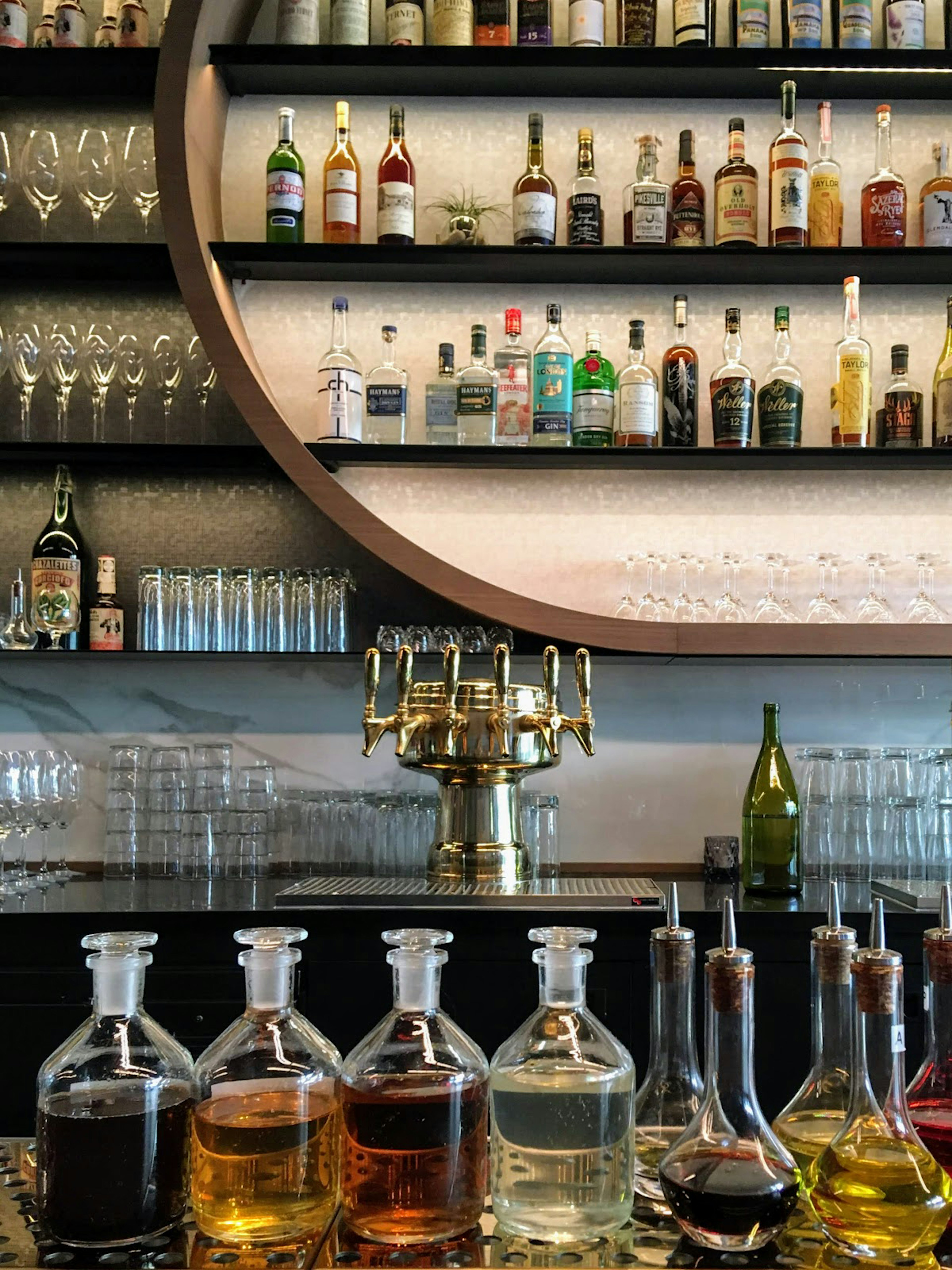 Glass bottles of liquor sit on a sleek and stylish modern bar with many other bottles lined up in the background