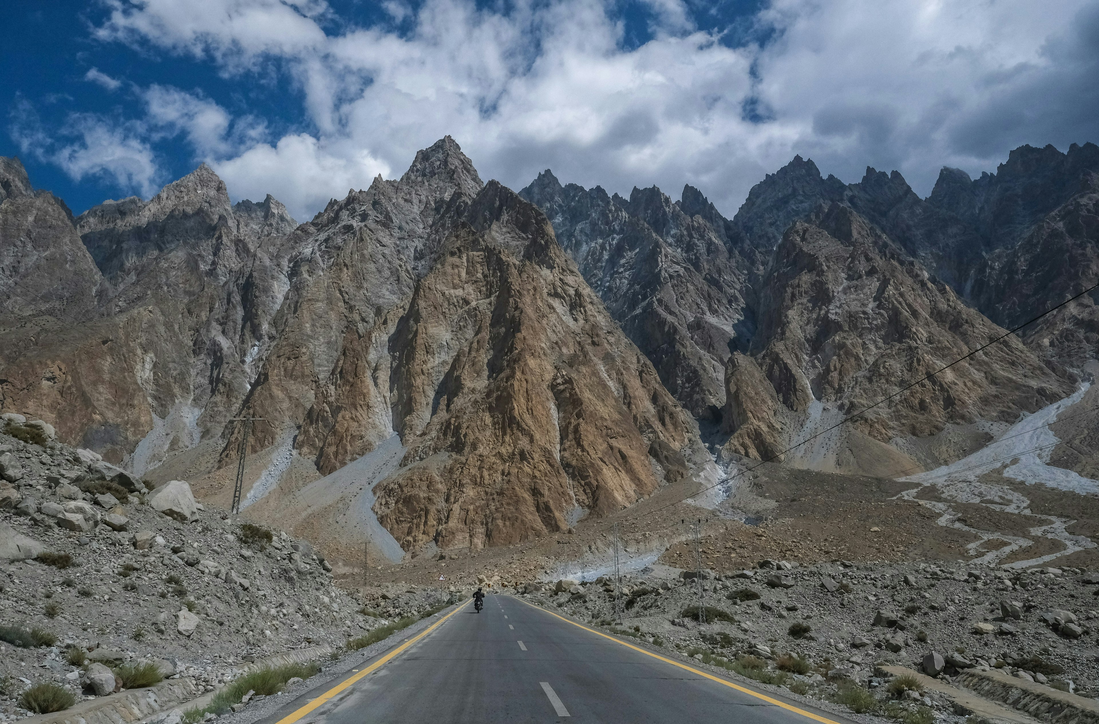 A faraway shot of a man driving a motorbike down a long, straight road. In the background, large mountains rise up towards the cloudy sky.