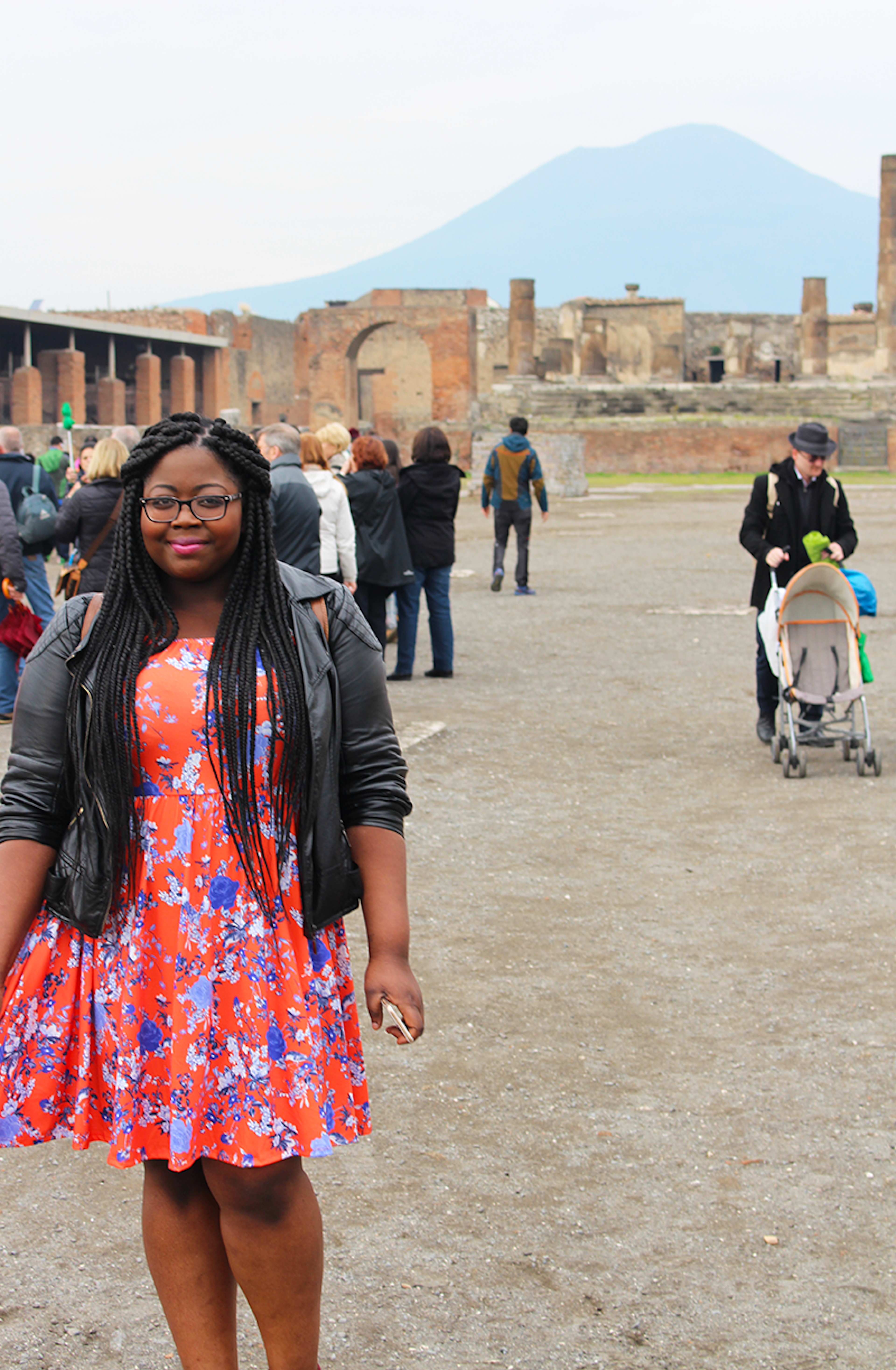 A young black woman poses in the foreground and some ruins in Pompeii are visible in the background. Other visitors mingle around; she is standing alone. Mount Vesuvius looms in the background of the photo. A black girl travelling through parts of Italy may experience racism.