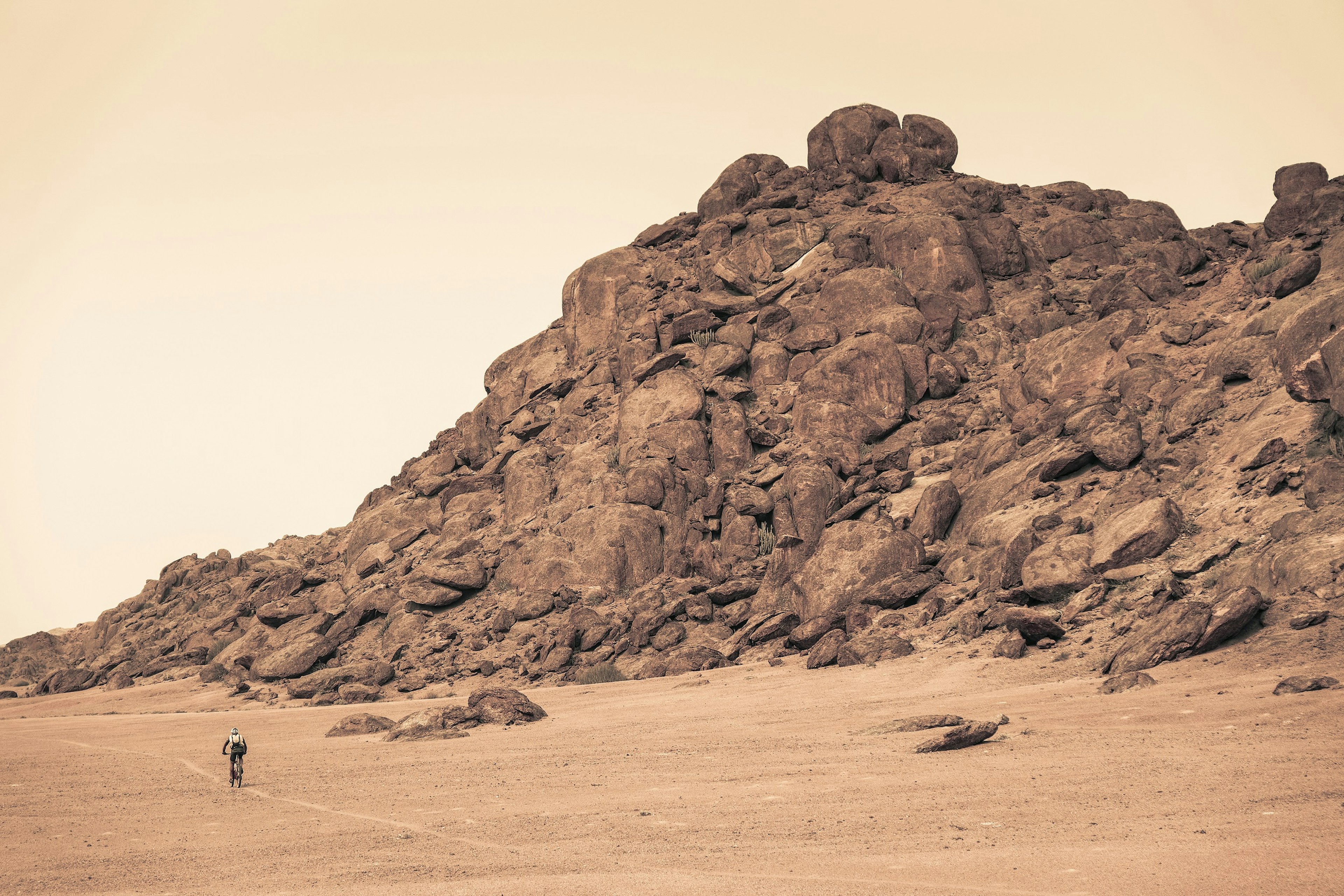 A lone mountain biker rides across a desert landscape past a large rock outcrop that towers above; everything is hued in brown tones.