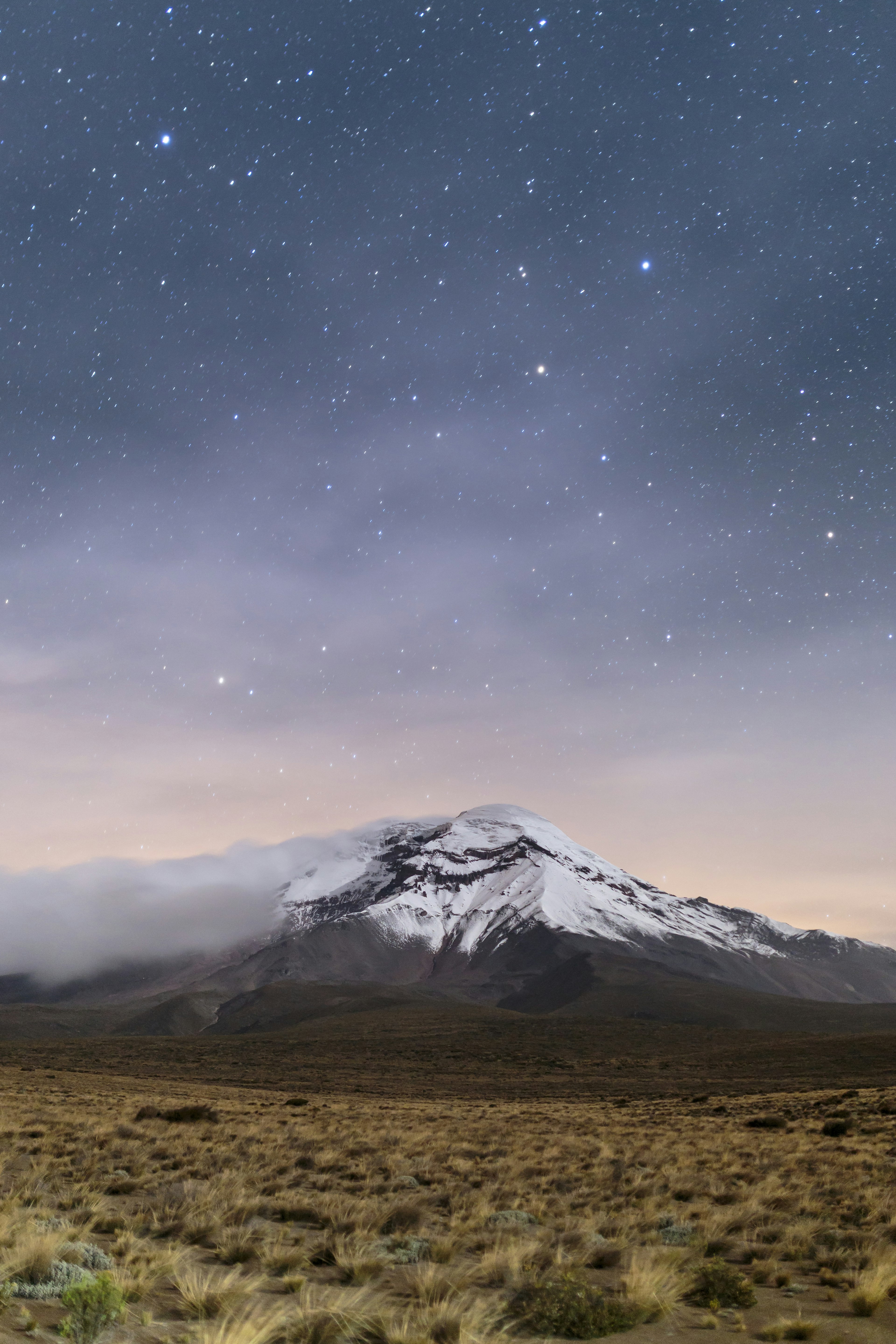 A snowcapped mountain sits at the edge of a grassland in Ecuador; starts permeate the clouds above.