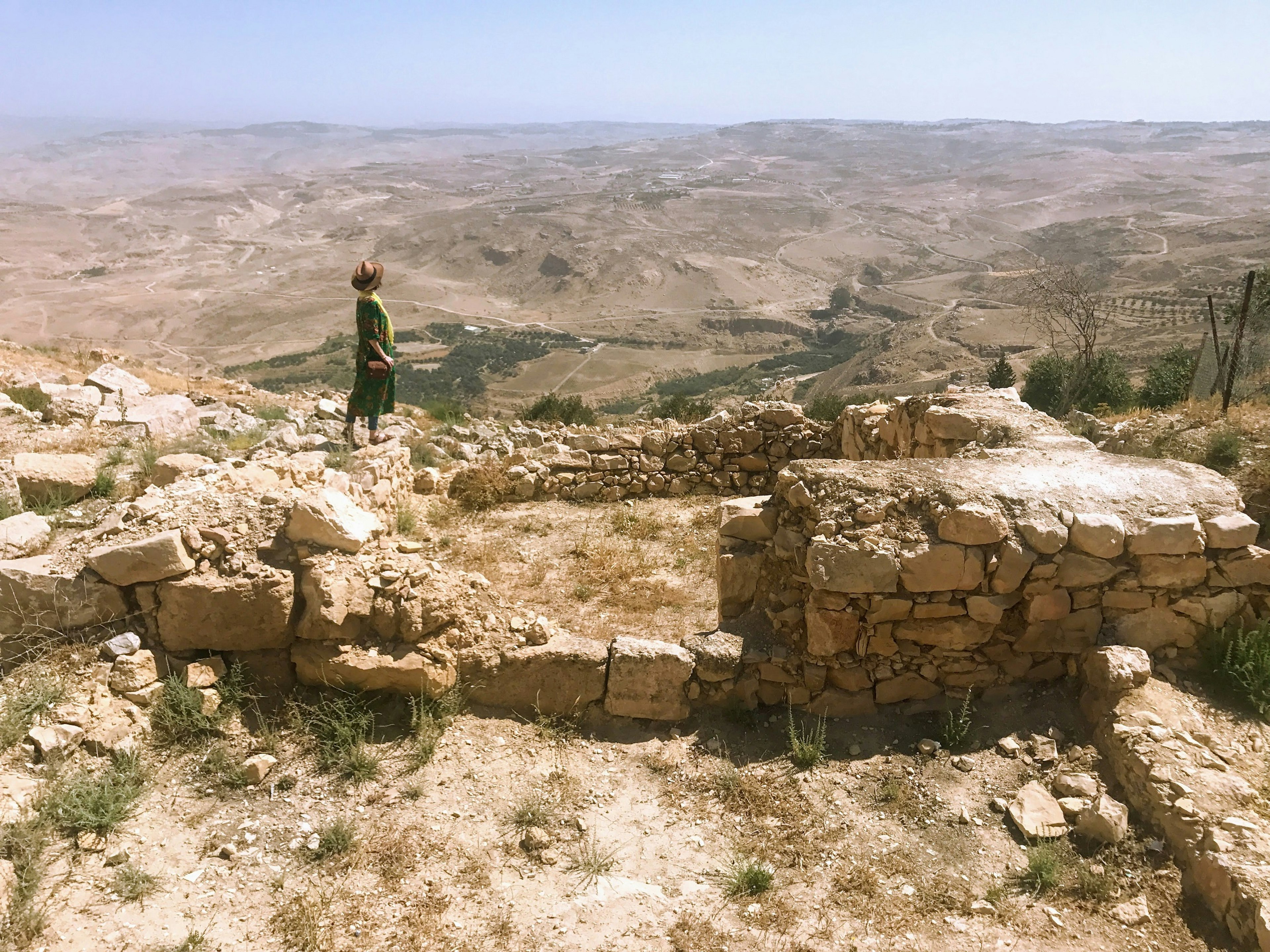 A woman in a green dress and straw hat stands on some ruins and looks over a vast and barren landscape of rolling hills.