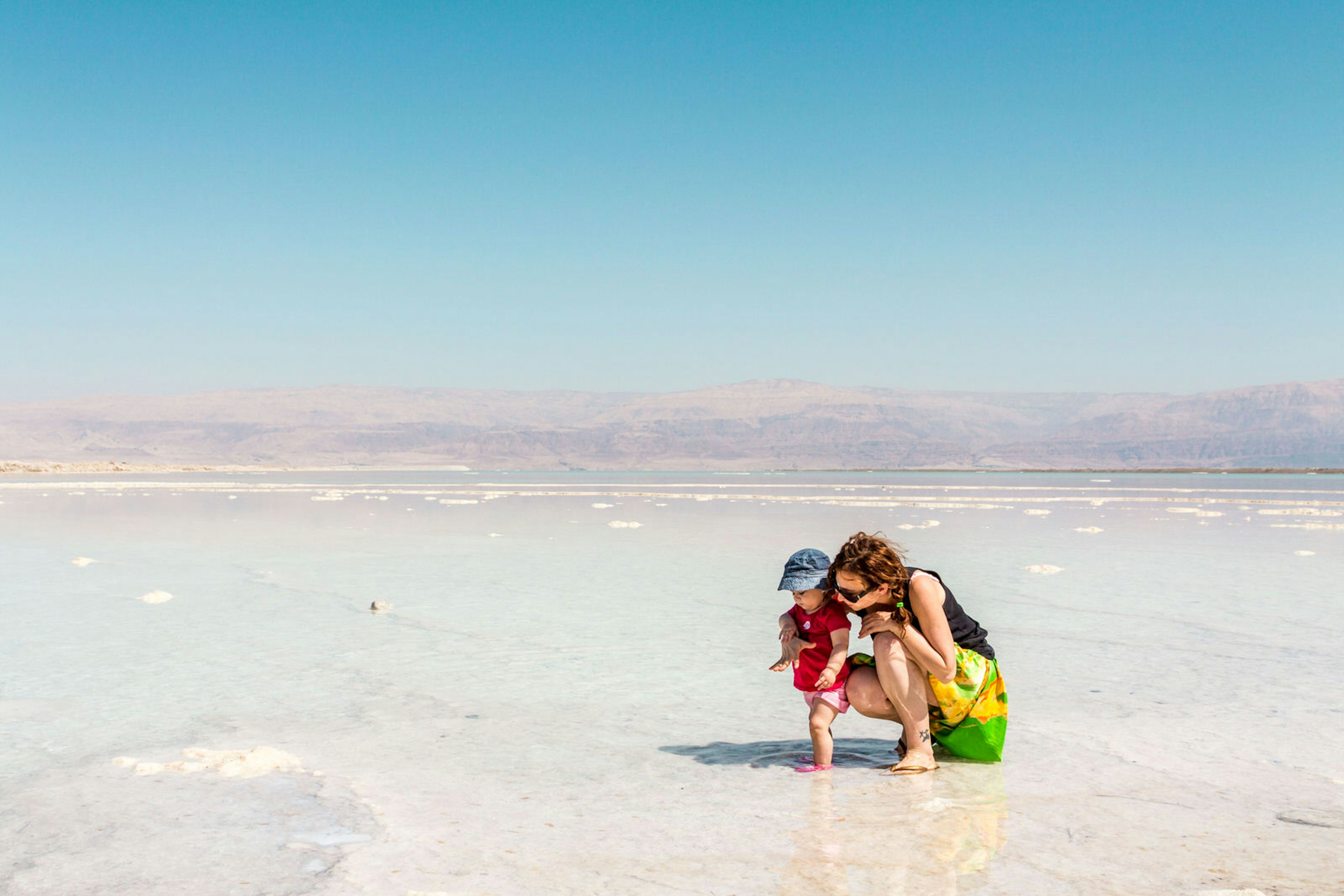 A mum squats down next to her hat-wearing child; both are standing in the glaringly white salt bed of the Dead Sea