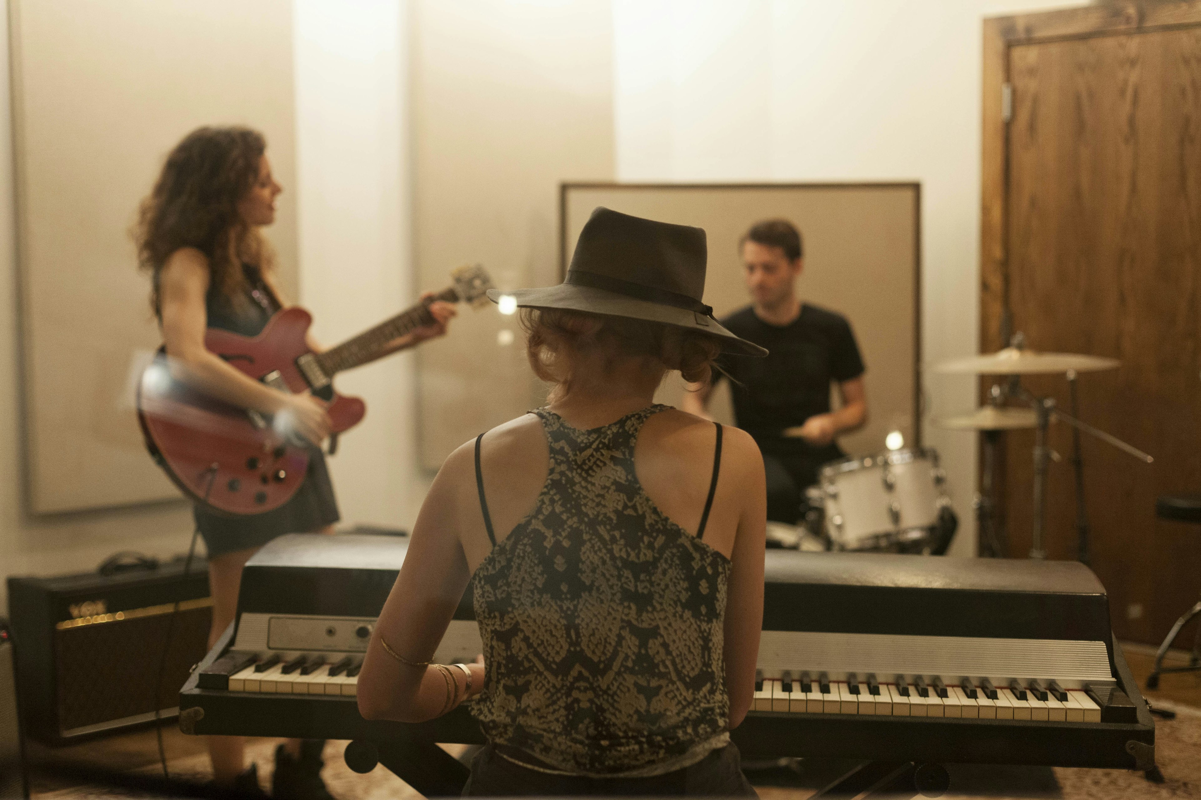 Musicians playing. A woman playing the piano has her back to the camera. To her left a woman is playing the guitar and in front of her is a man playing the drums