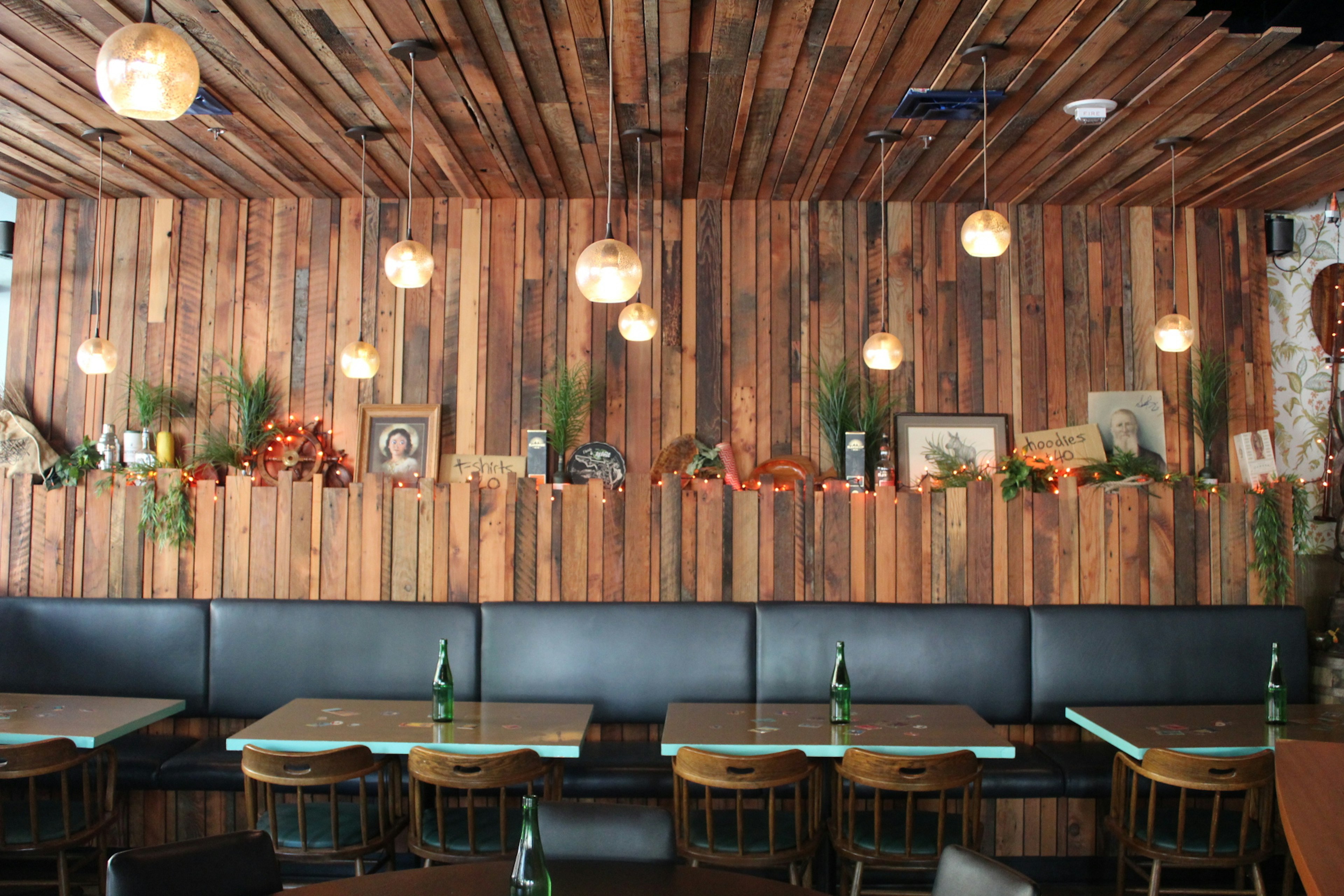 The interior of Navy Strength, a tiki bar in Seattle, is lined with worn wood panels from the top of the black banquet seating to the ceiling, which is also lined. White round lamps hang from the ceiling, and a shelf displays framed art, tchotkes, sea grass, and other ephemera